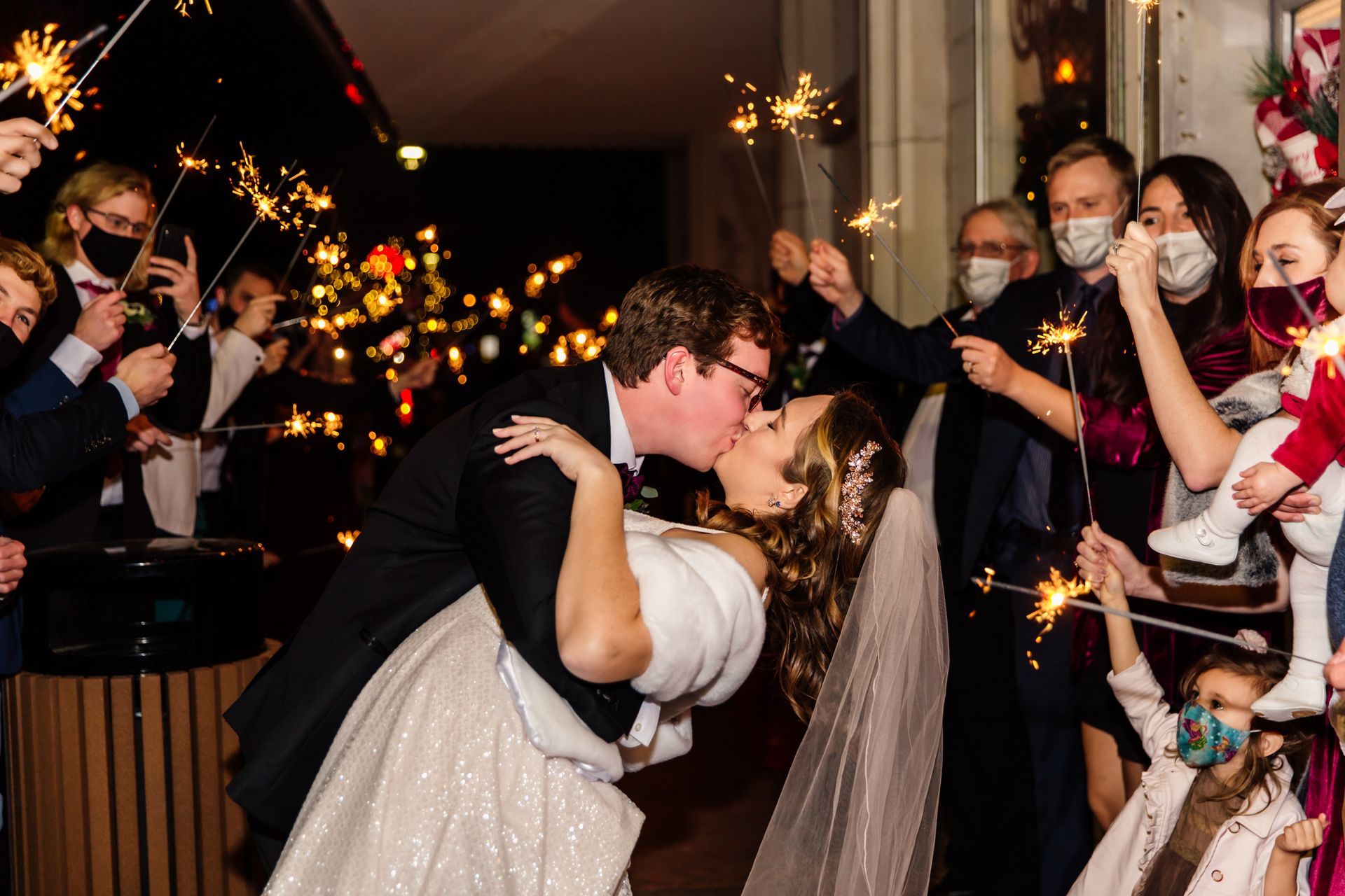 Couple kissing with sparklers held by masked wedding guests outside at night.