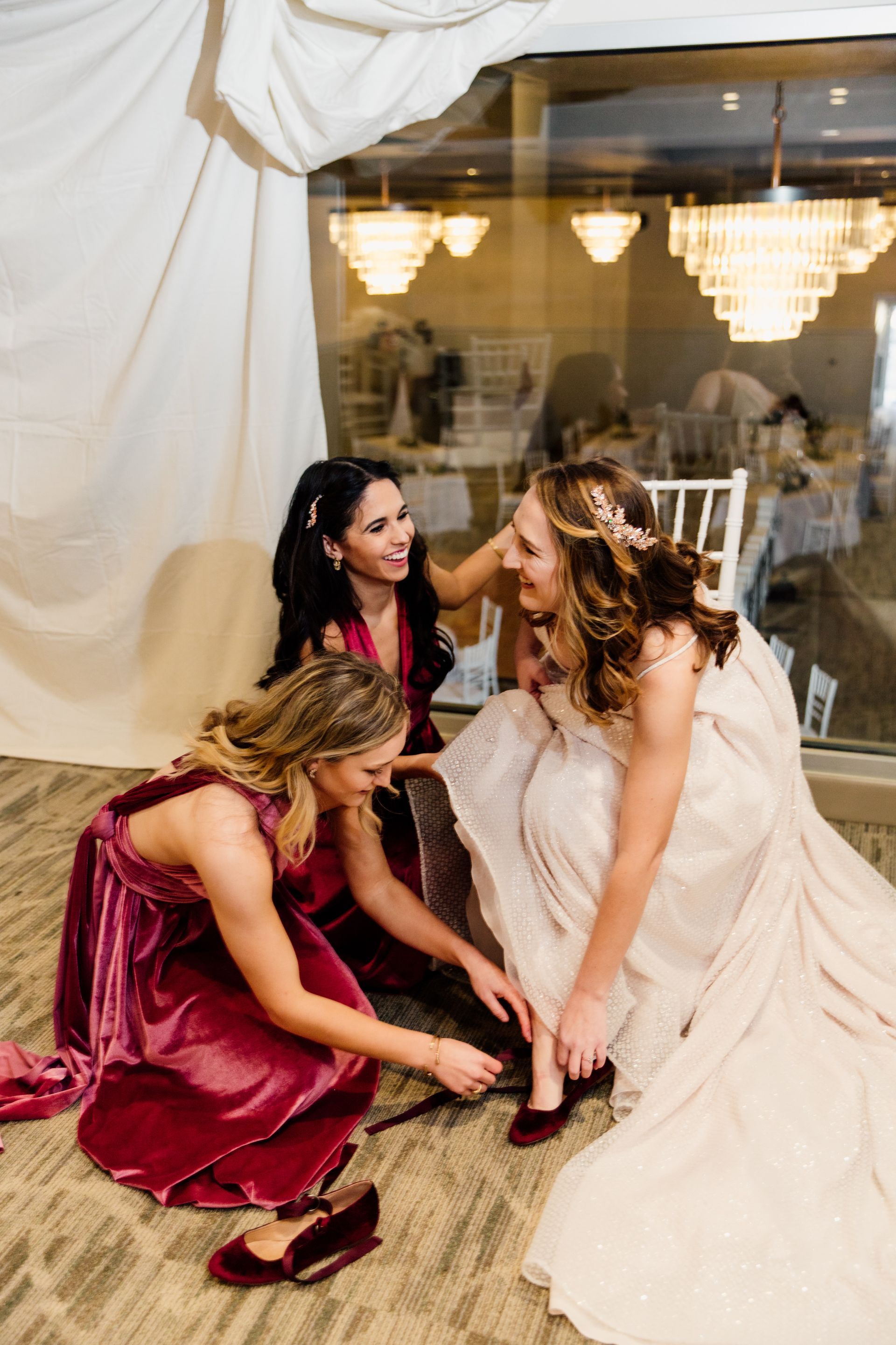Bridesmaids help the bride put on shoes before a wedding ceremony. Red and gold hues, indoor setting.