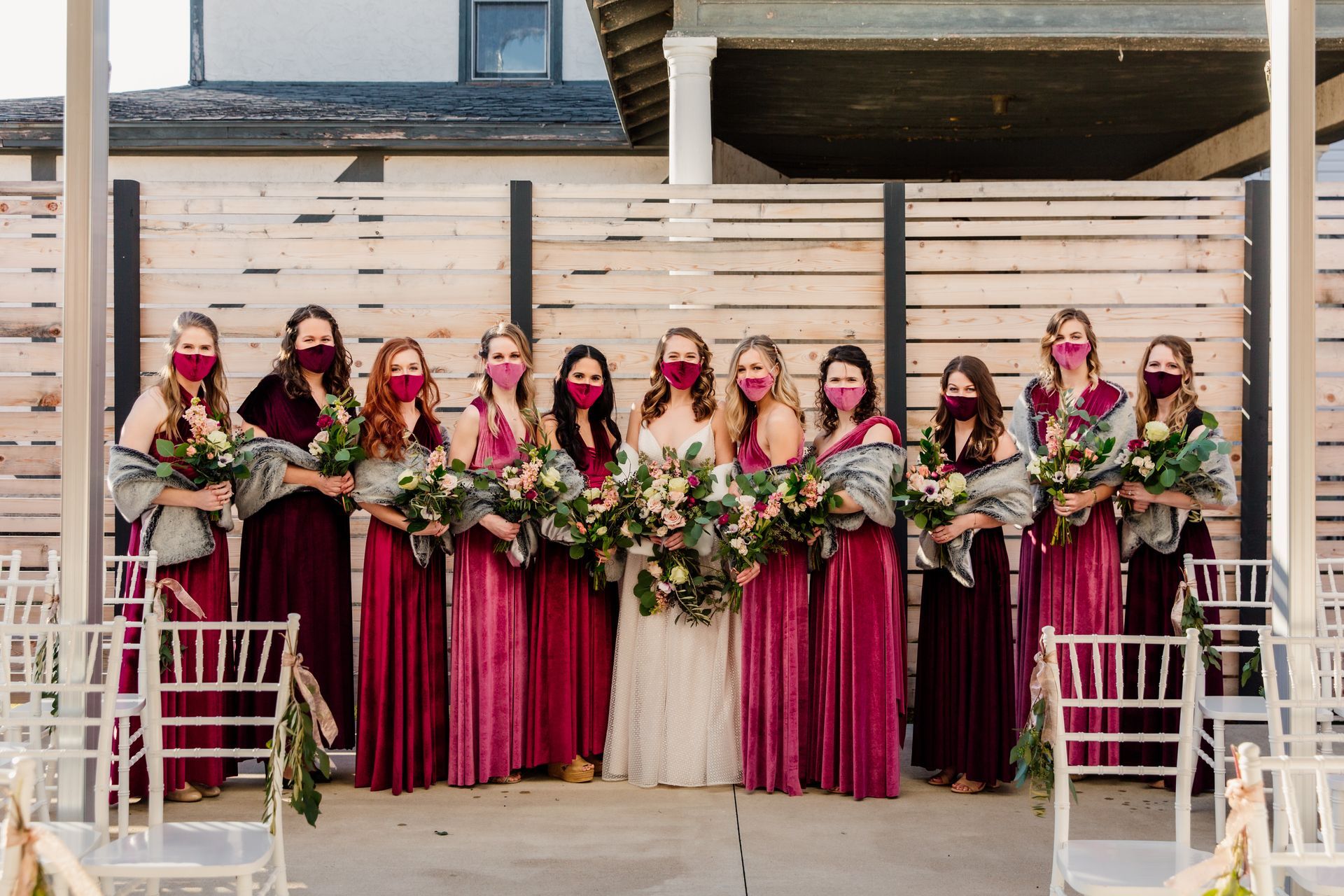 Bridesmaids with masks in burgundy dresses, holding bouquets, pose with the bride in front of a wood fence.