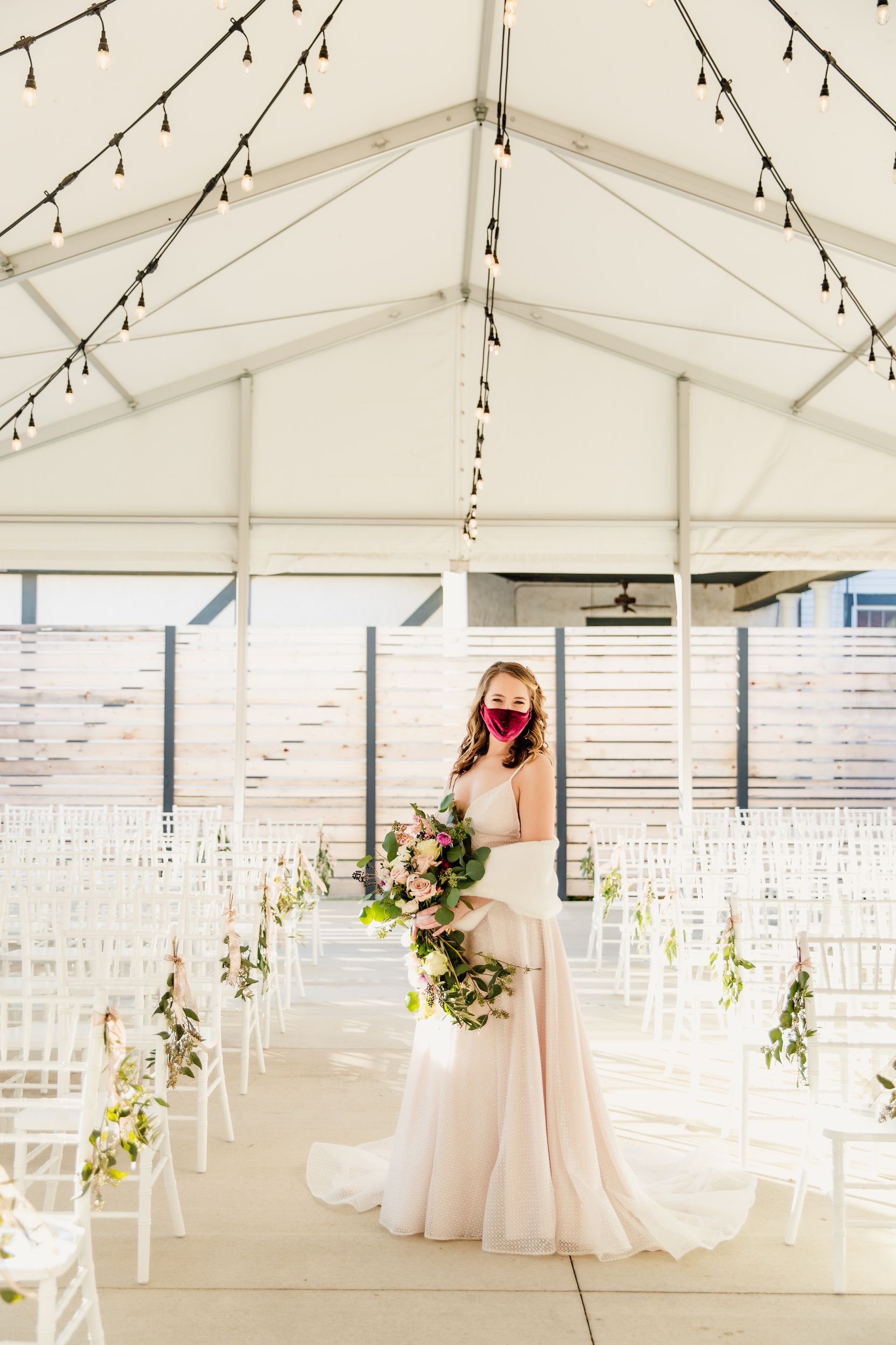 Bride wearing a mask holding flowers, standing under a tent with string lights, wedding ceremony setting.