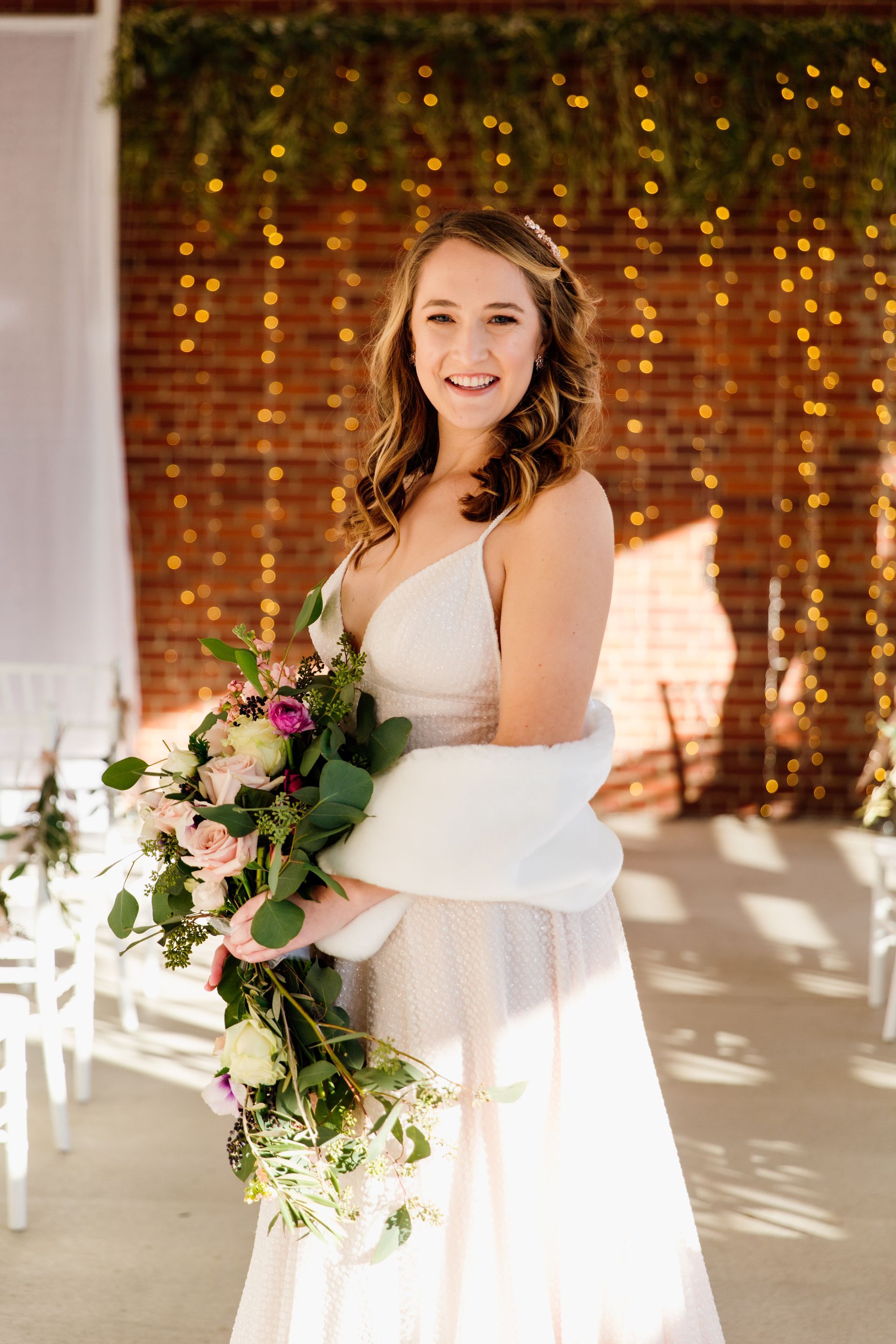 Bride smiles, holding a bouquet, wearing a white dress and shawl, standing in front of a brick wall decorated with lights.