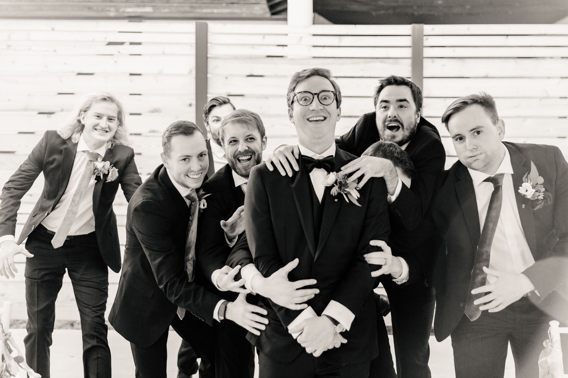 Groomsmen playfully surround a man in a tuxedo, smiling at the camera, in front of a wooden wall.