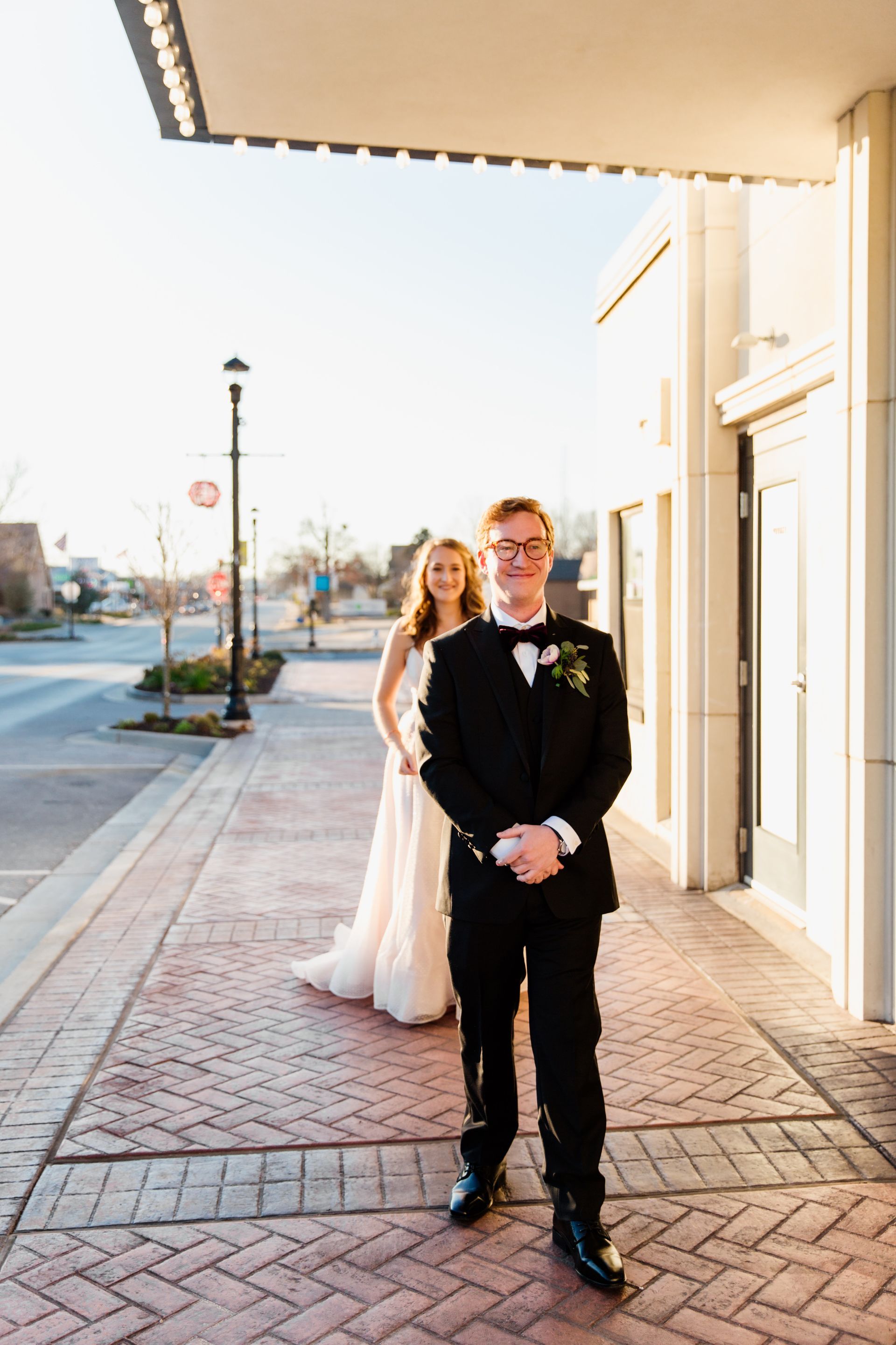 Groom in black tuxedo smiles, bride in white dress behind him, standing on brick sidewalk near building.