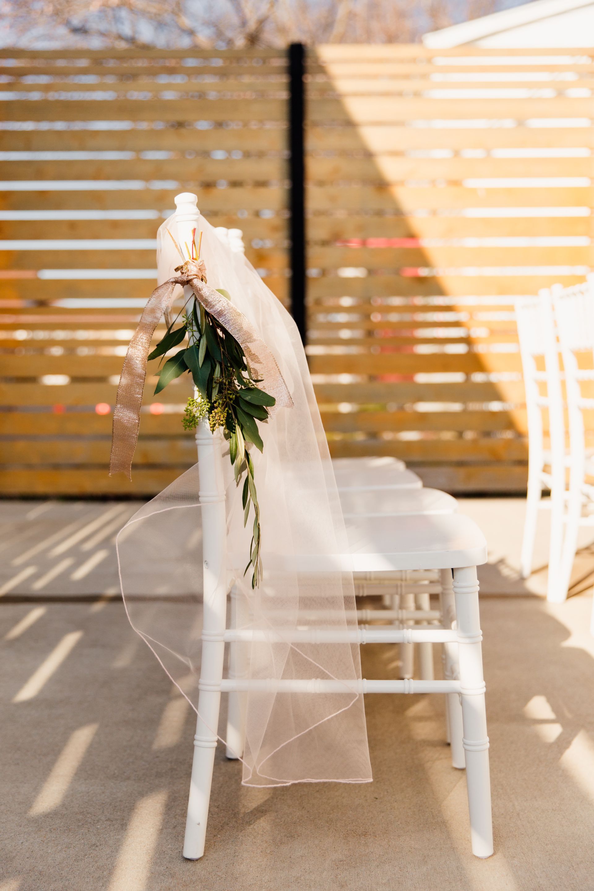 White chair decorated with veil and greenery, set on a patio. Wooden fence in background.