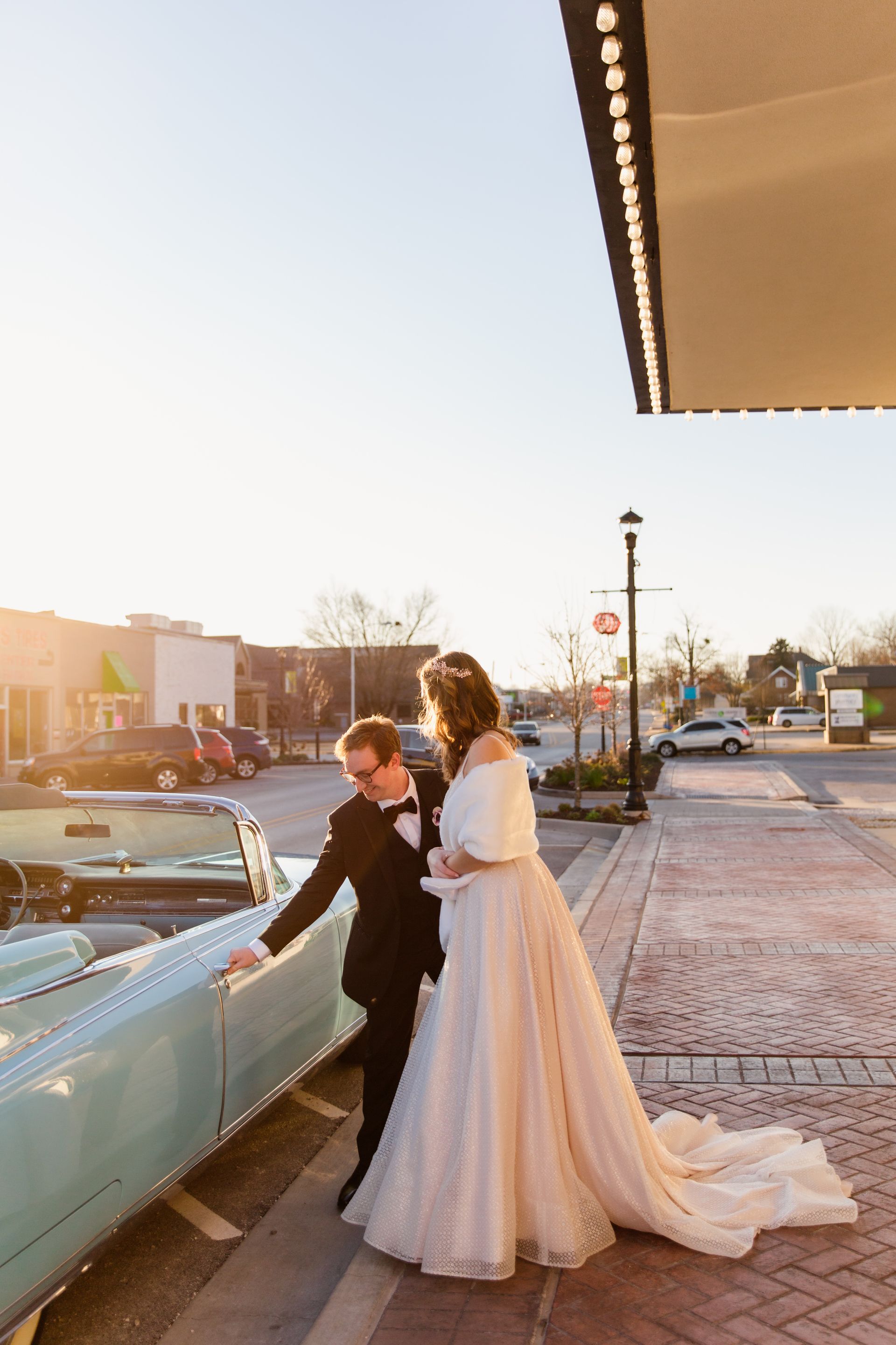 Bride and groom by a vintage teal car on a city street at sunset.