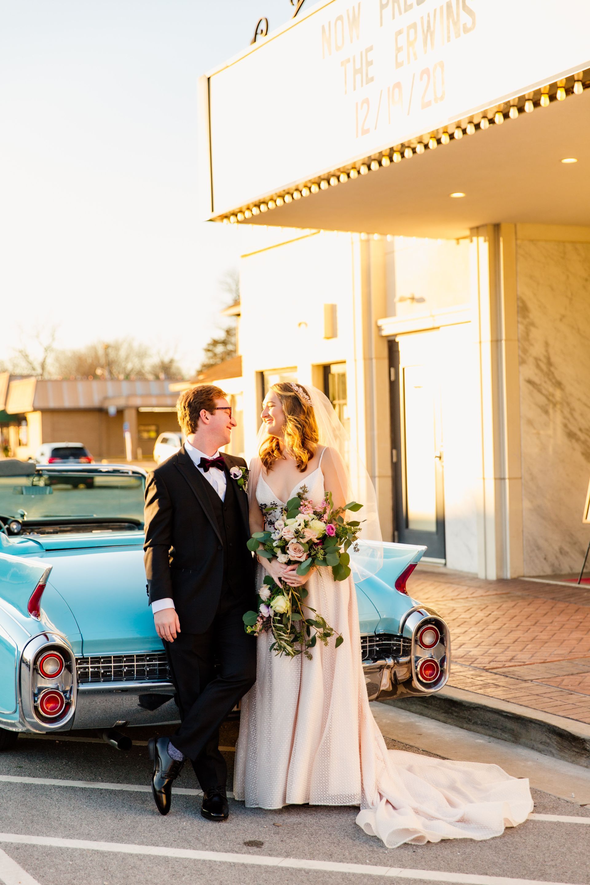 Bride and groom by a blue vintage car in front of a theater. Golden sunlight bathes them as they smile.