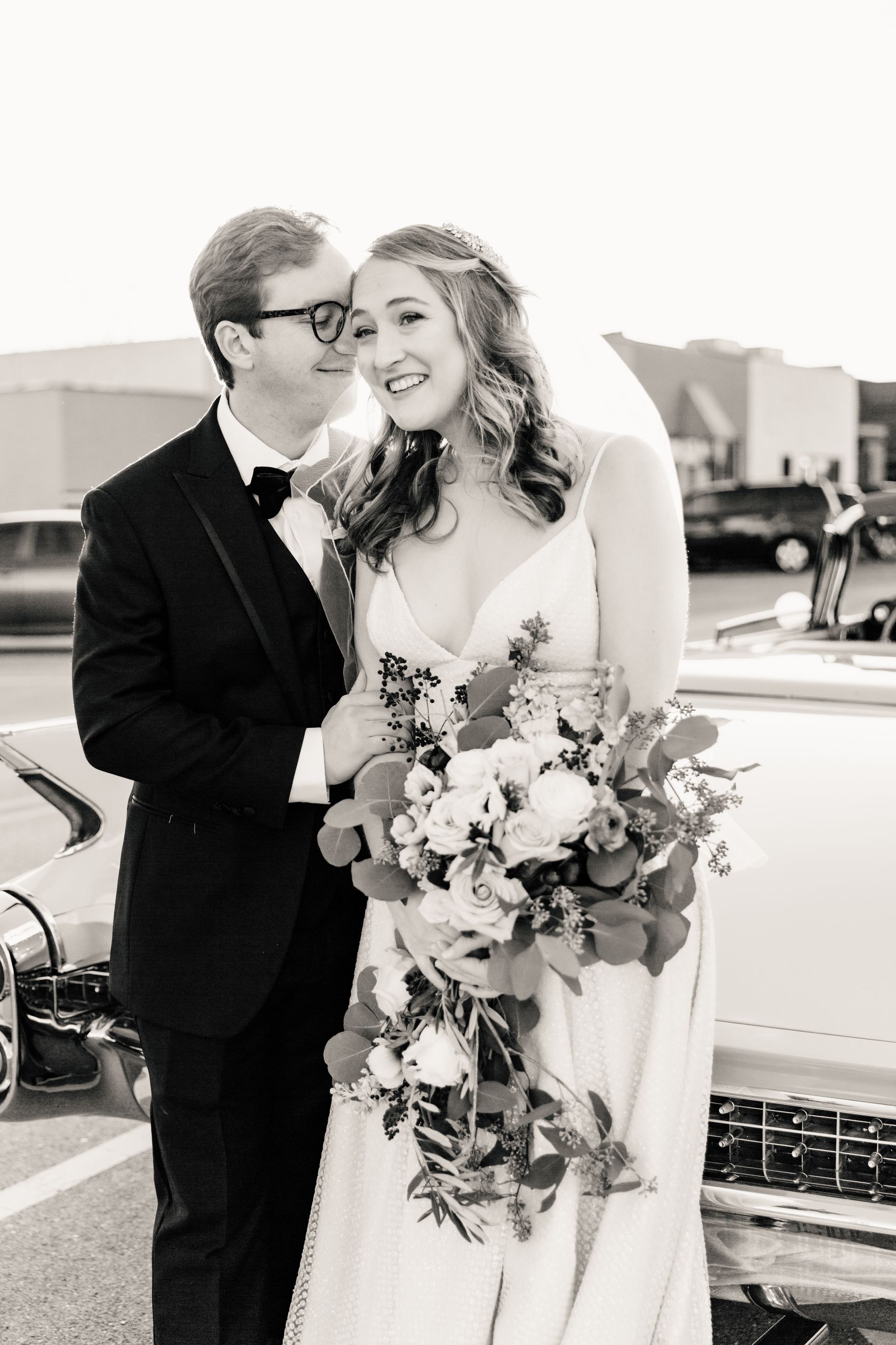 Bride and groom in formal attire embrace outdoors, smiling. Bride holds a bouquet, groom wears a bow tie.