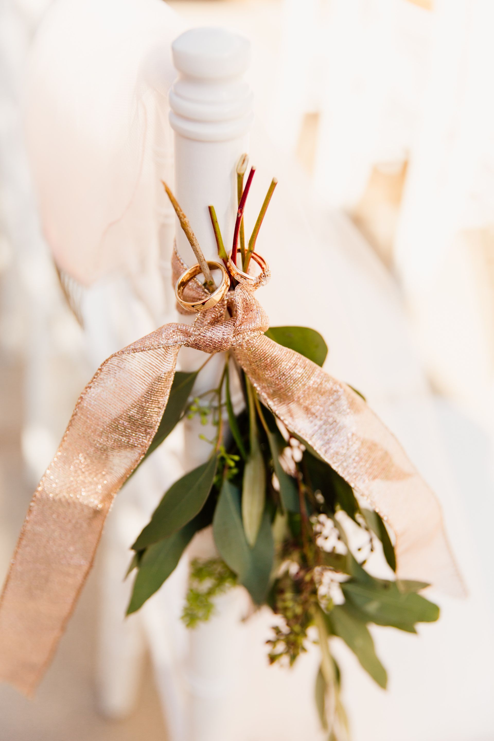 White chair decorated with a gold sequin bow, greenery, and small twigs for a wedding.