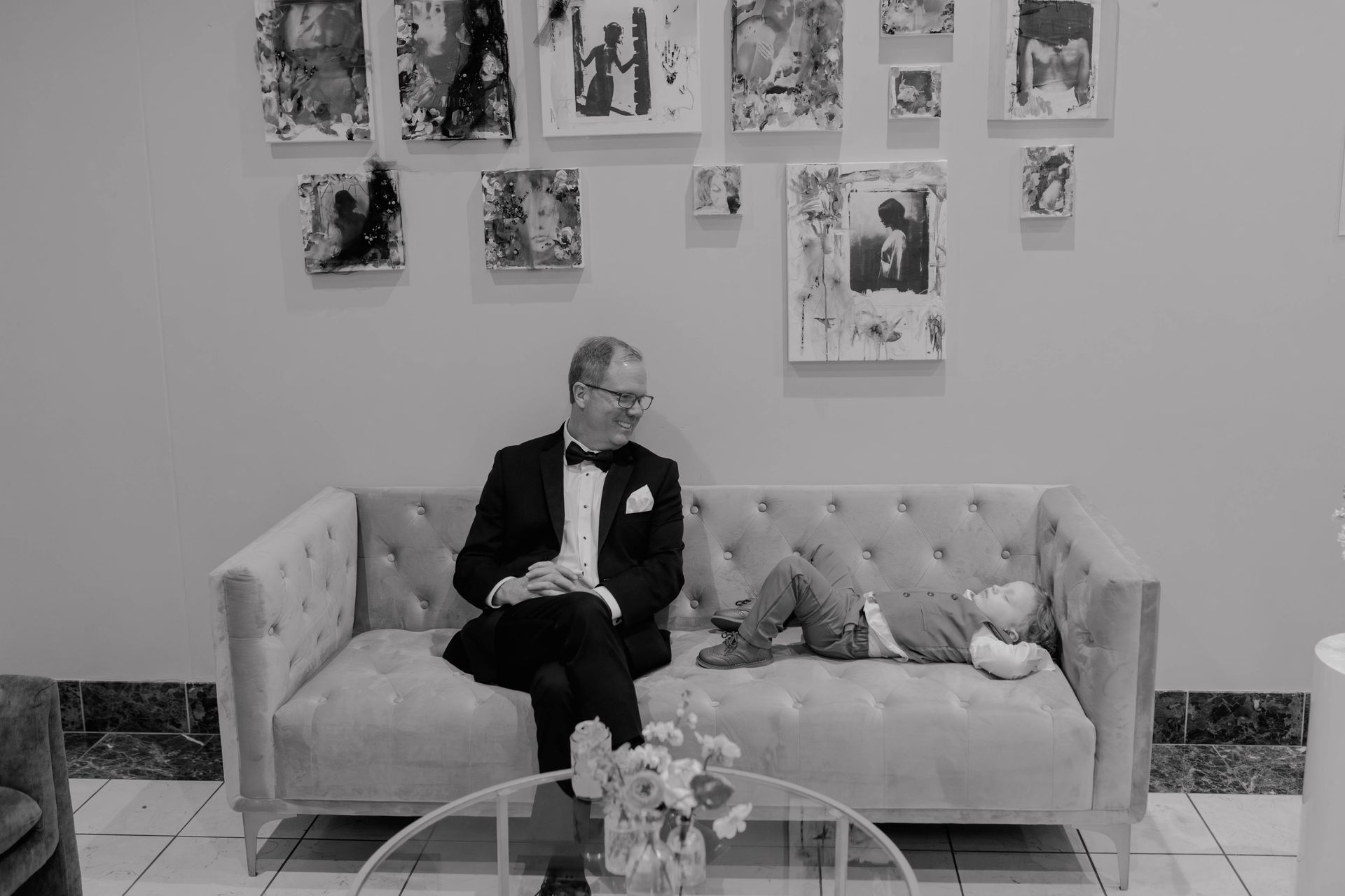 Man in tuxedo sits on a sofa, looking at a child lying down. Paintings hang on the wall.