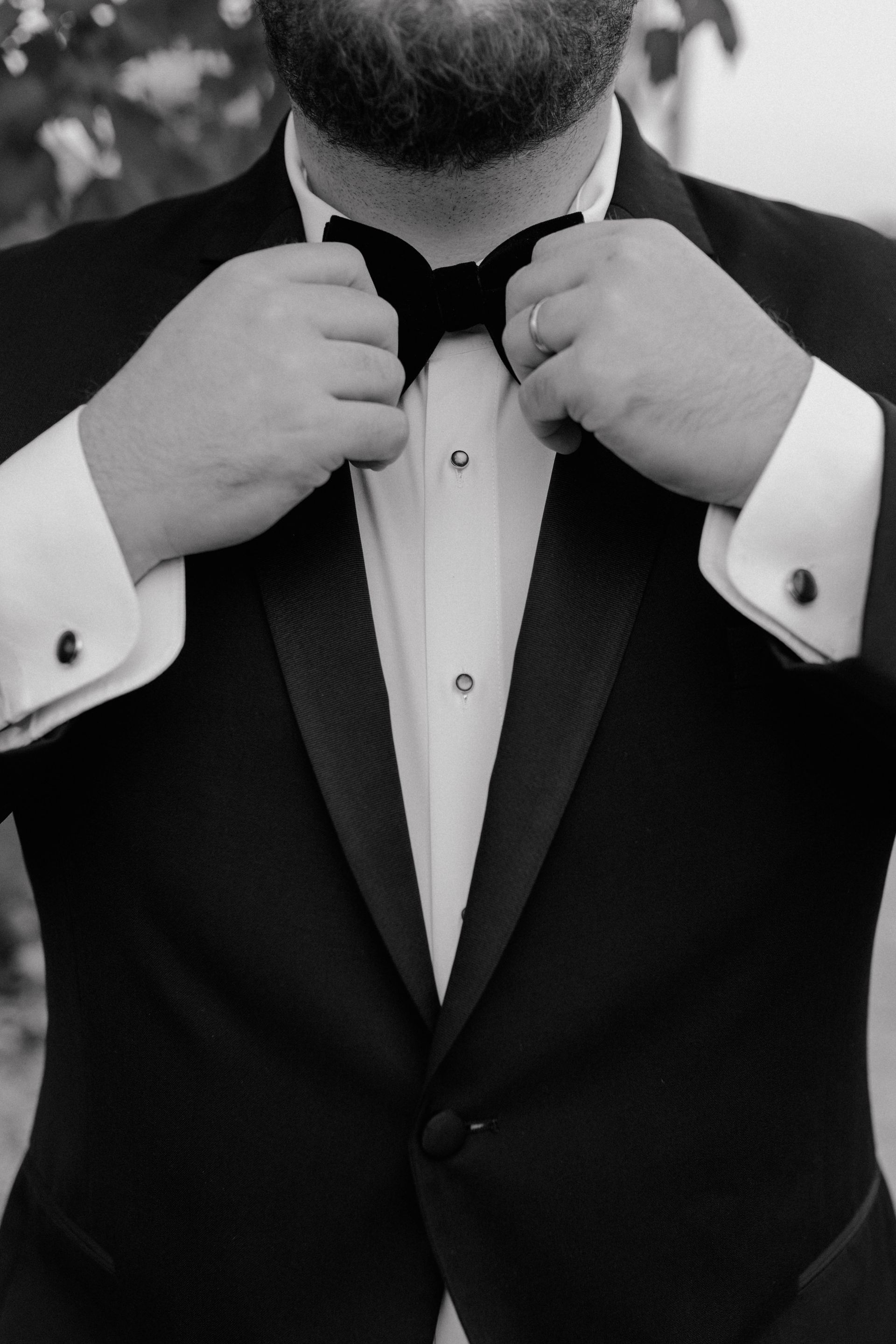 Man adjusting his black bow tie, wearing a tuxedo with a white shirt and cuff links.