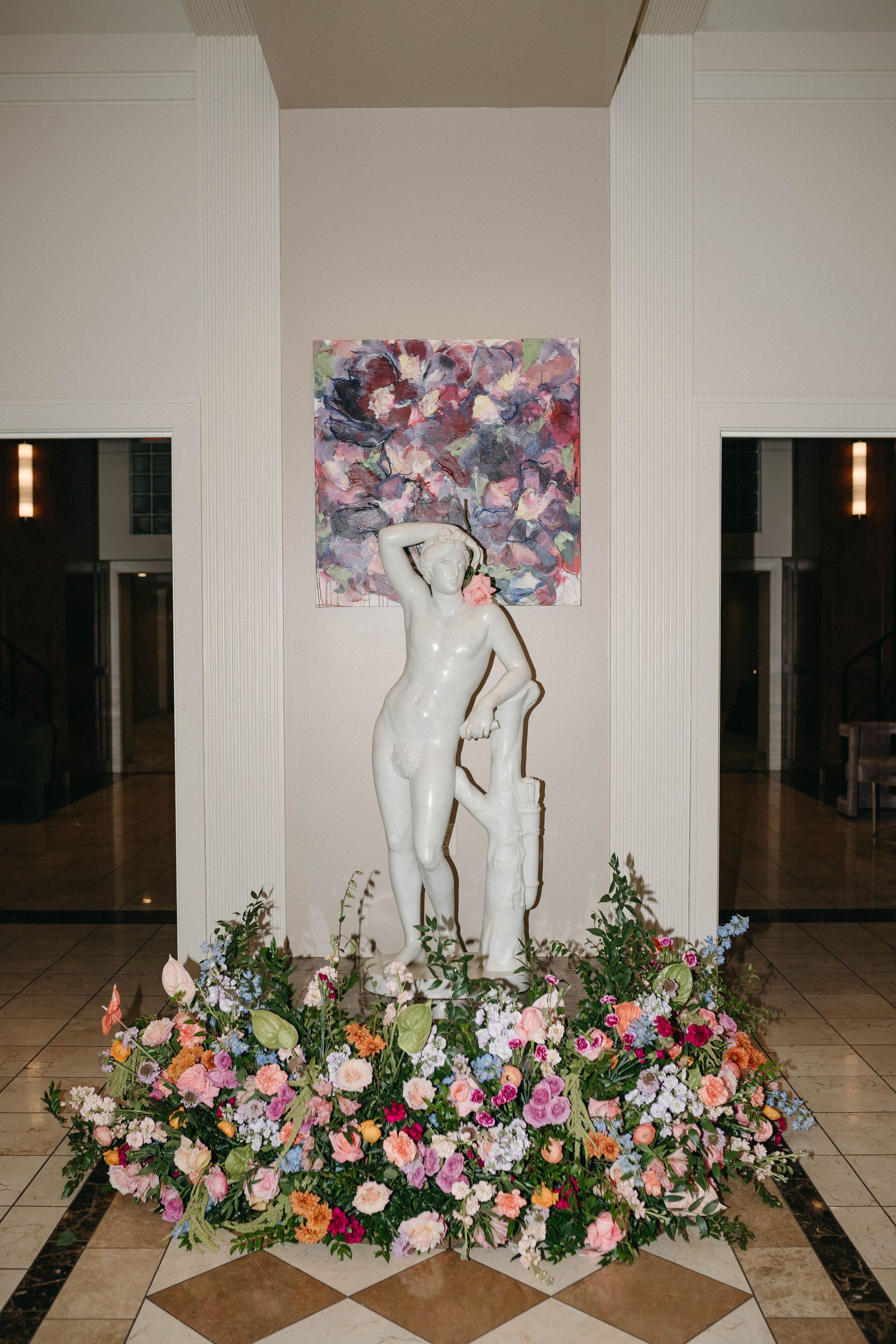White statue with floral arrangement in front of a colorful abstract painting on a wall in a building's entrance hall.