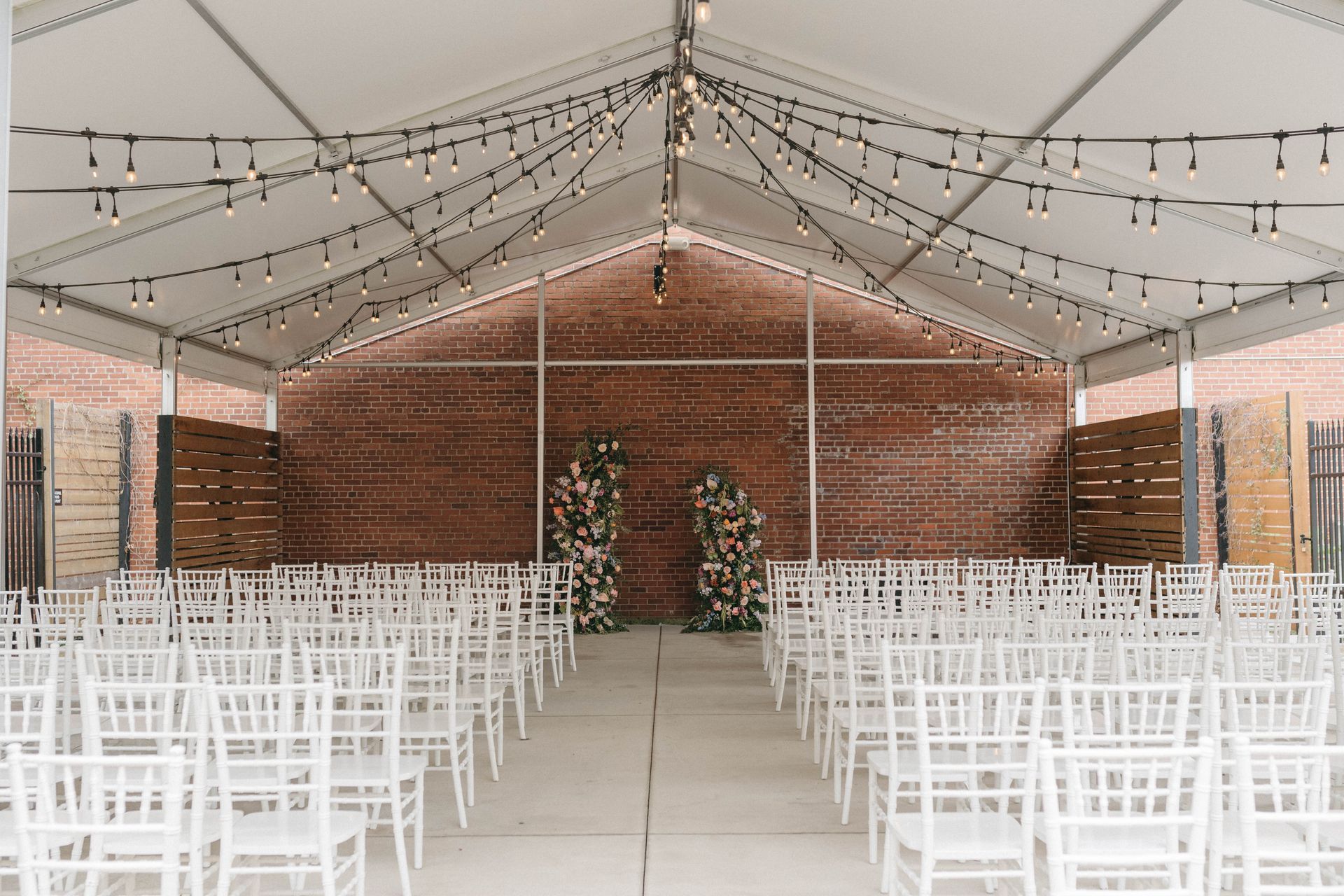 Rows of white chairs face a floral-decorated altar inside a white tent lit with string lights.