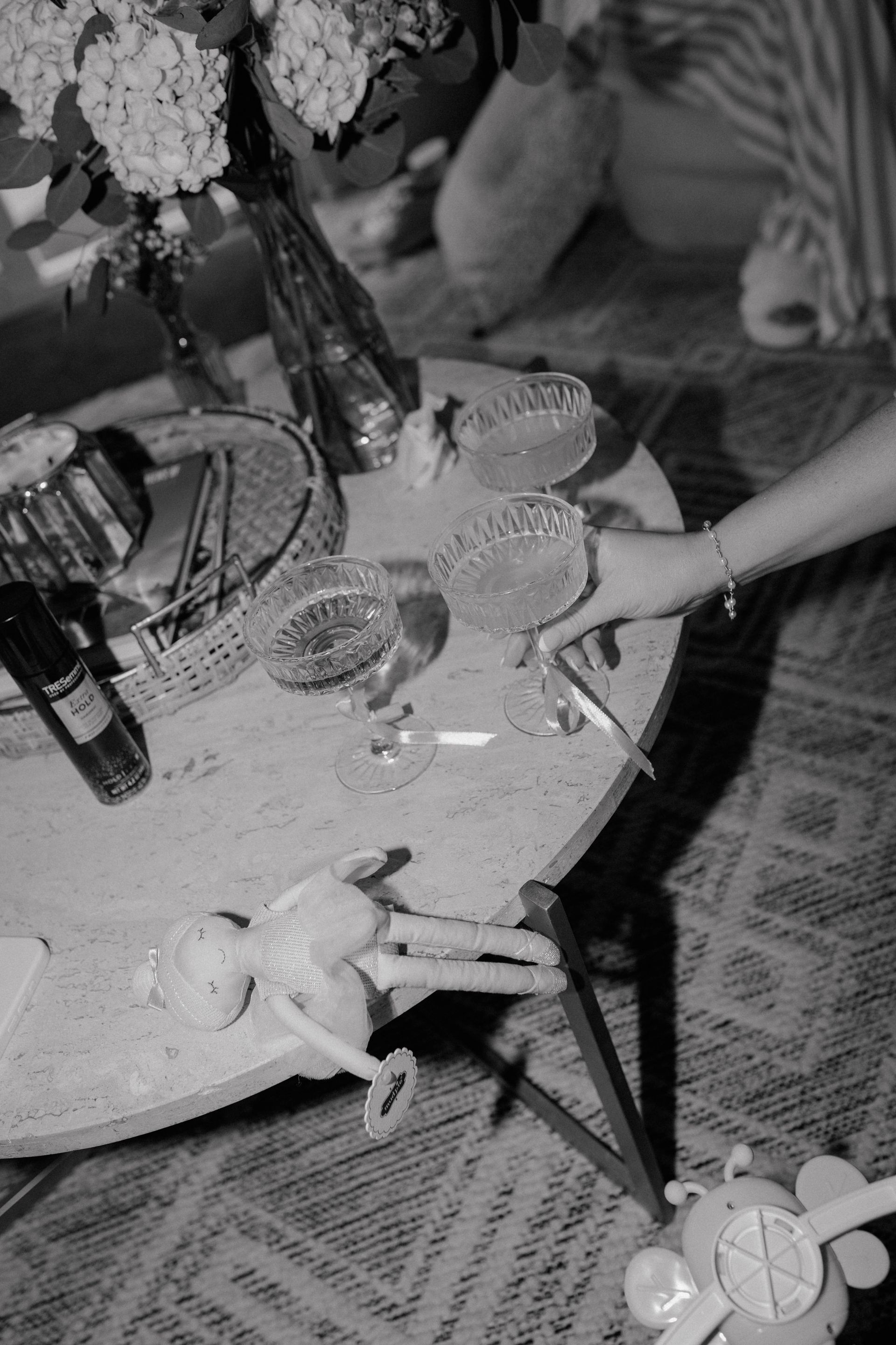 Black and white, round table with drinks, flowers, and a doll. Hand reaches for a cigarette.