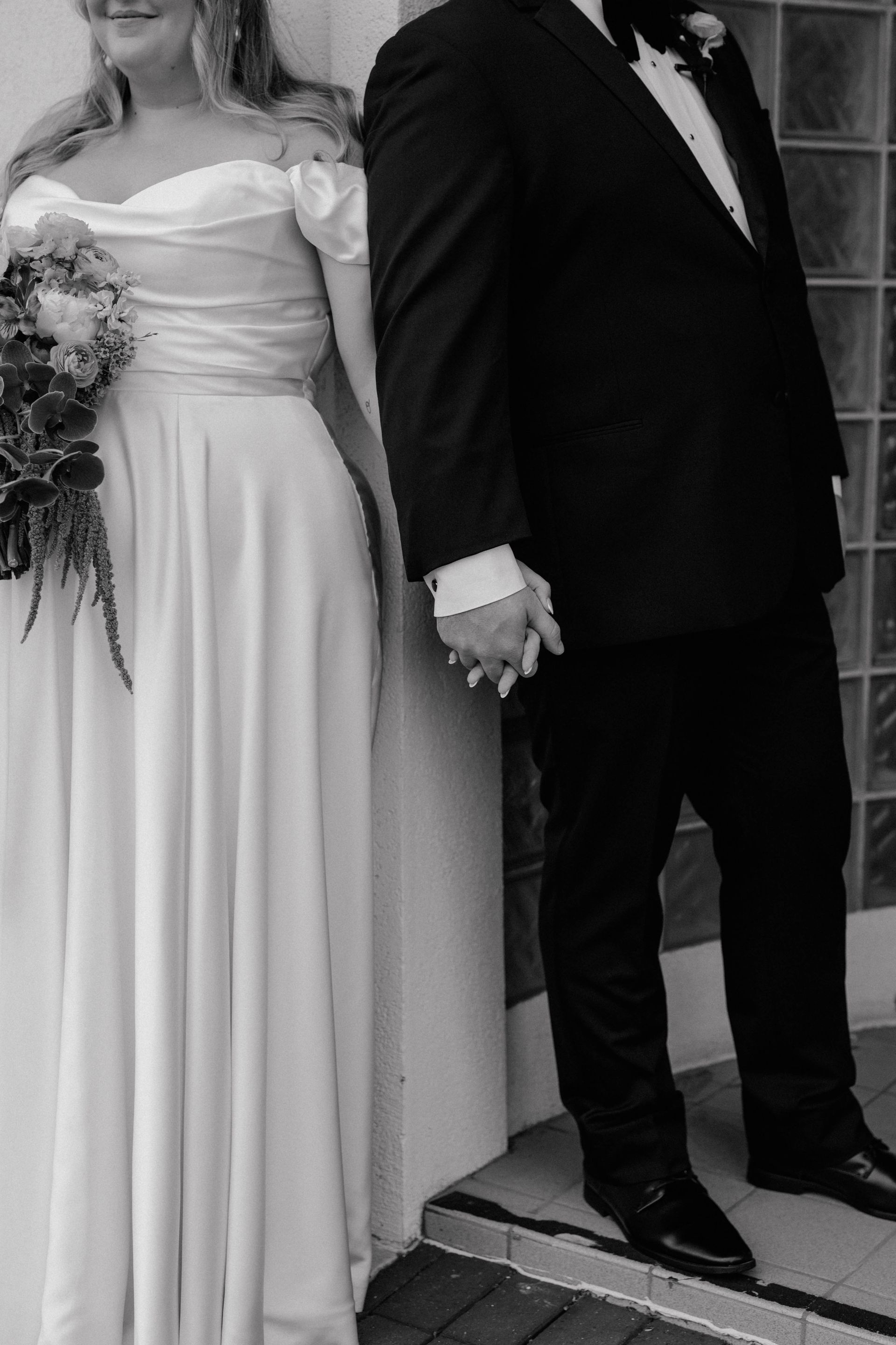 Bride and groom holding hands, bride in off-white dress, groom in black tuxedo, by a building.