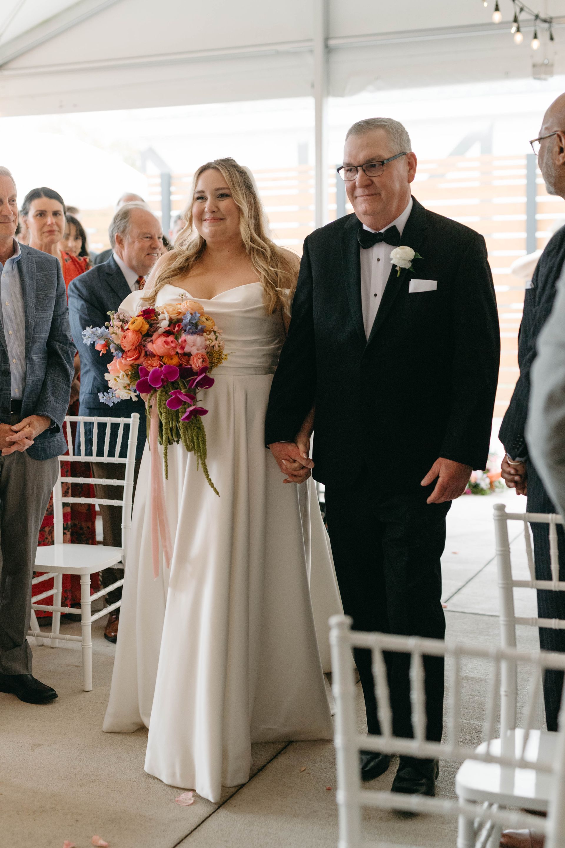 Bride and her father walk down the aisle at an outdoor wedding. She holds a colorful bouquet.