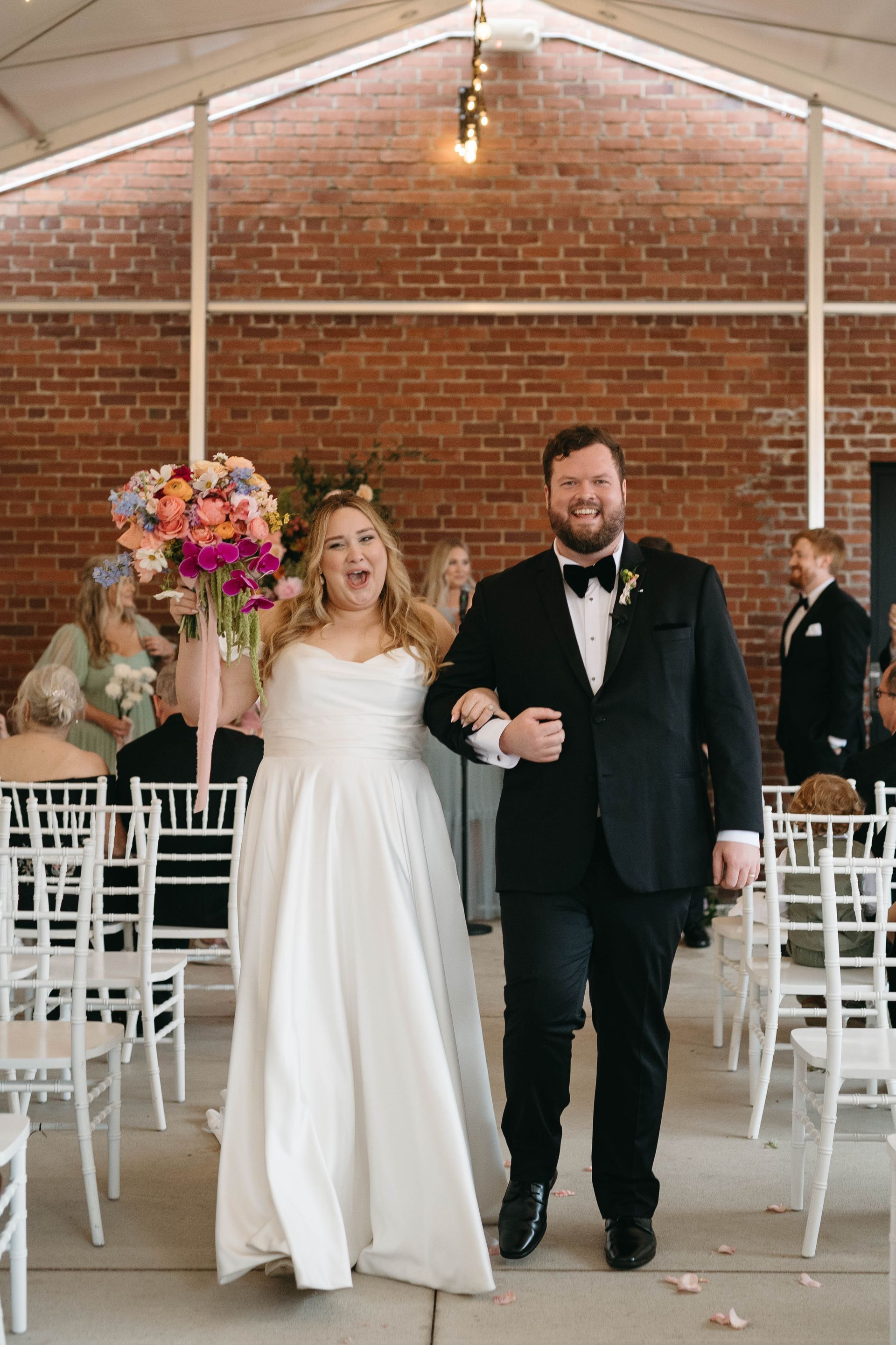 Newlyweds exiting a brick-walled venue, bride holding colorful bouquet and smiling, groom in tuxedo.