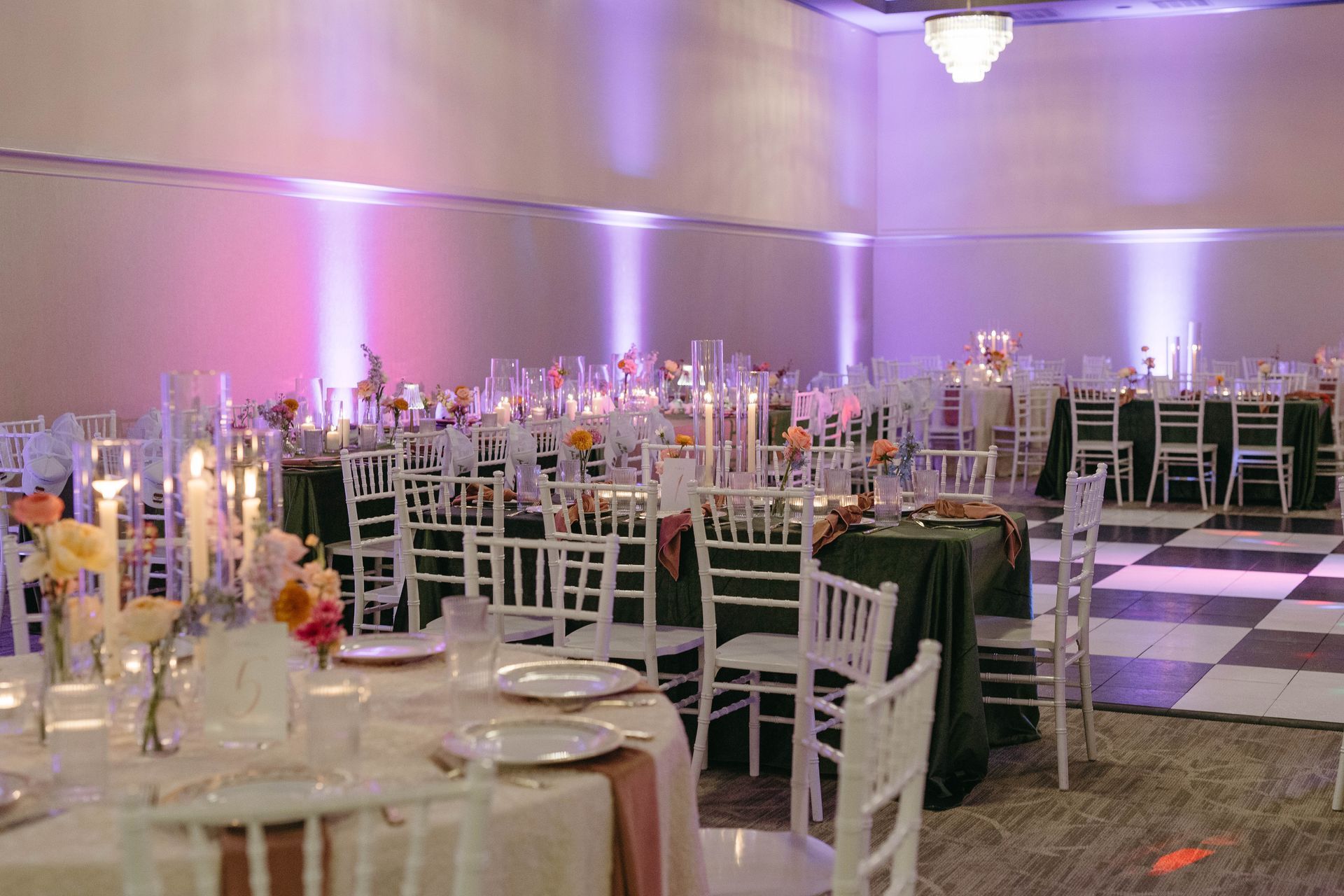 Banquet hall with decorated tables, white chairs, and dark green tablecloths, lit with purple and white lights.