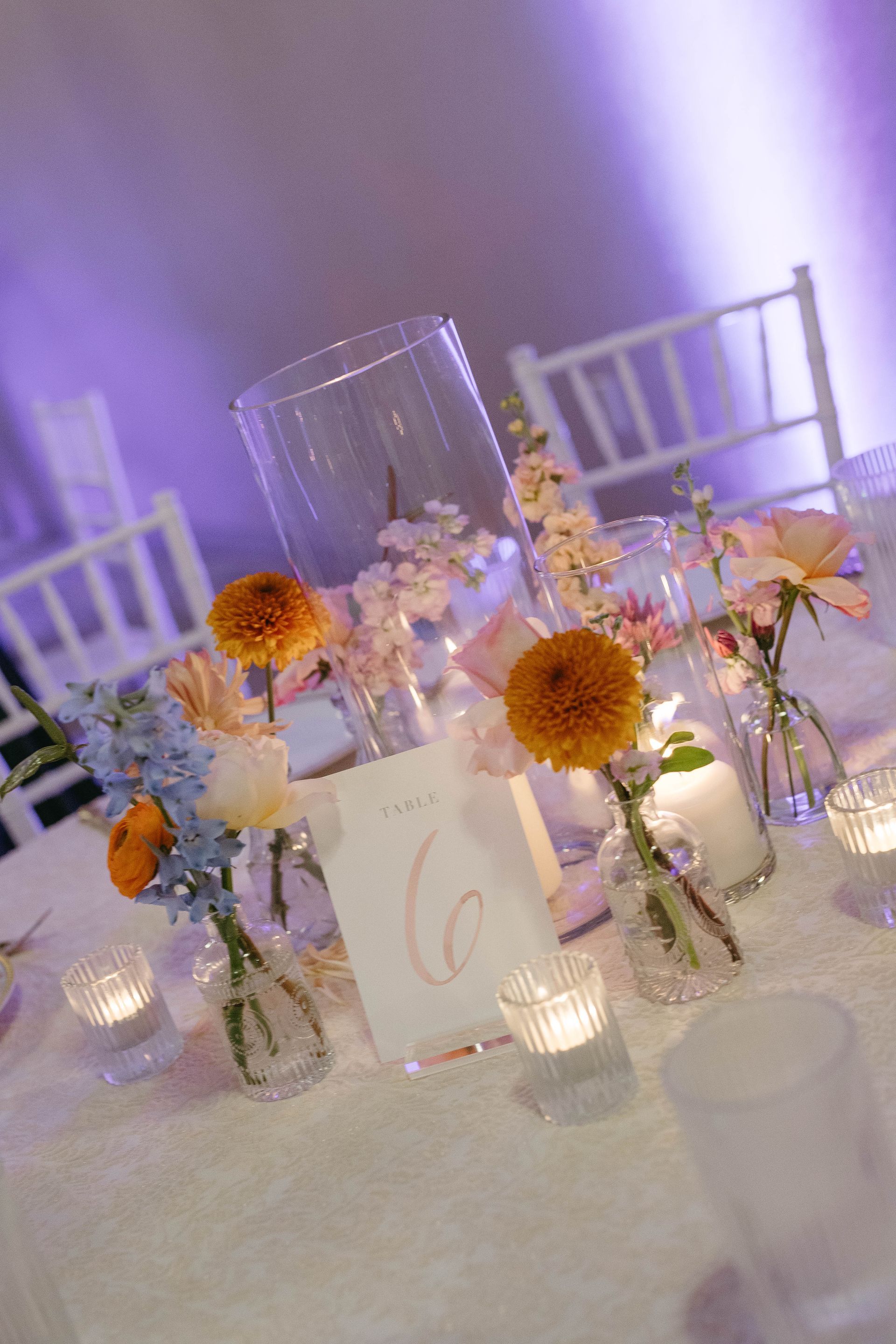 Table setting with flowers, candles, and a table number, under purple lighting.