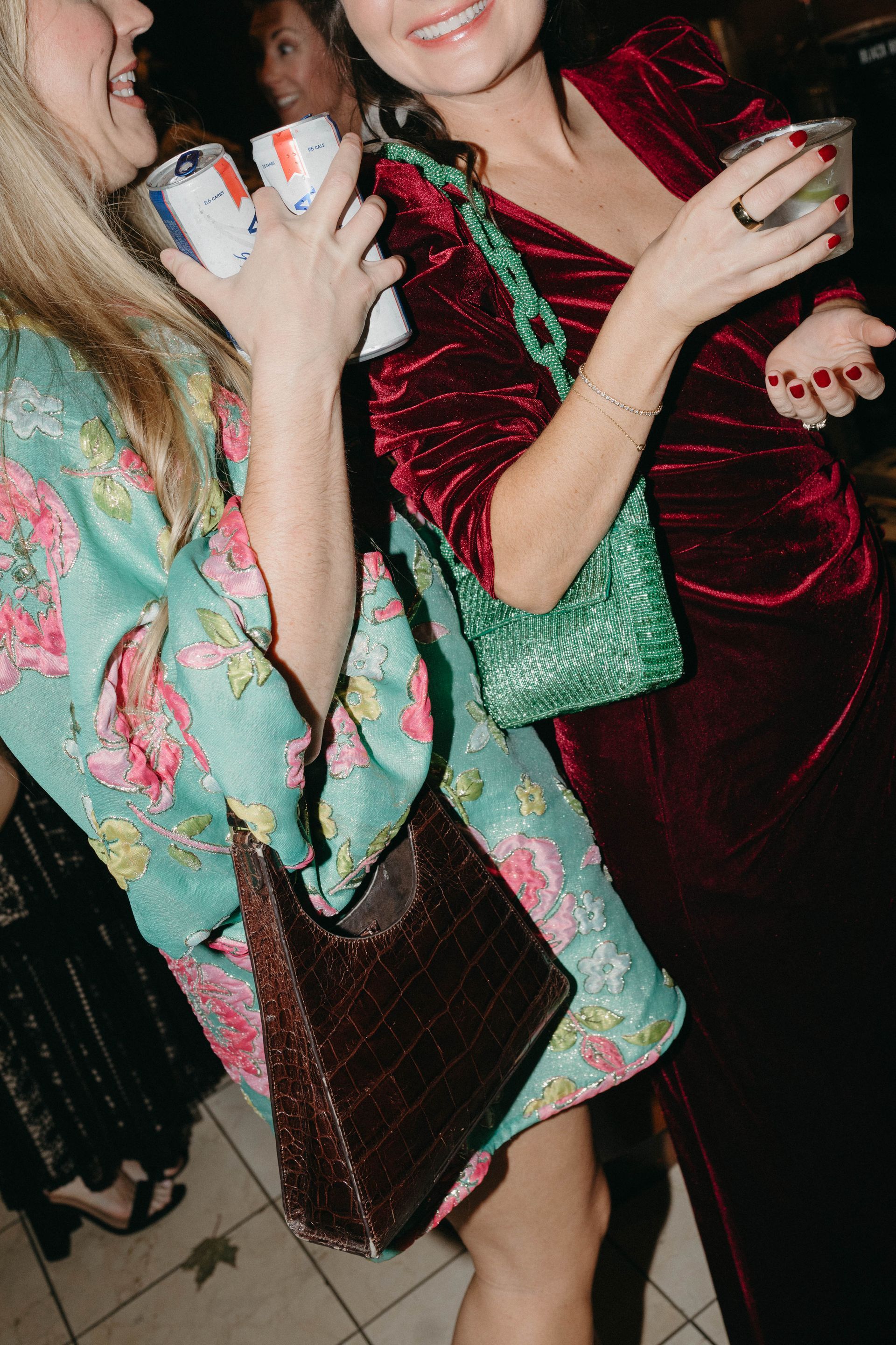 Two women laughing, one in floral dress, other in red velvet, holding drinks at a party.