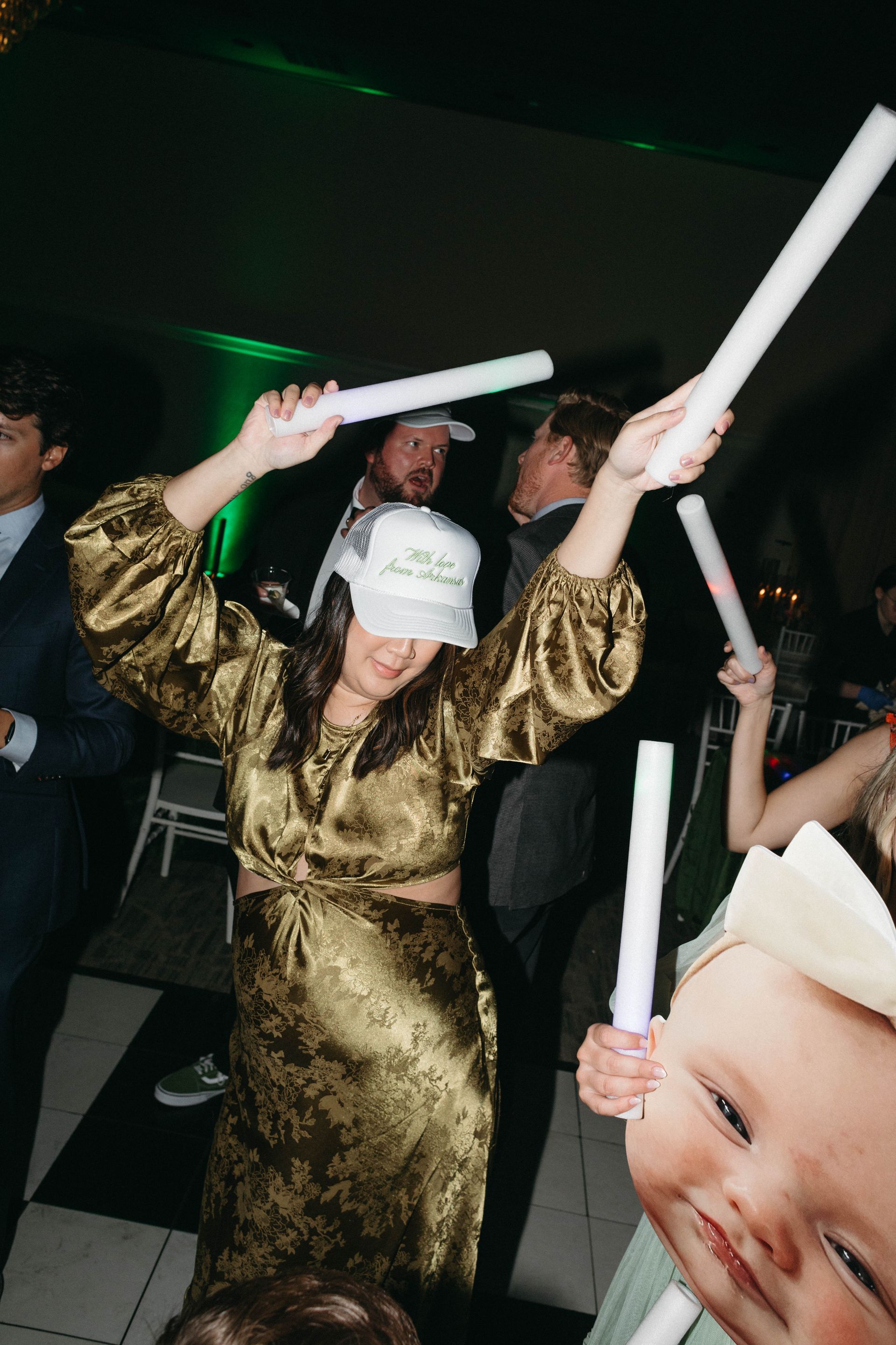 Woman in gold dress dancing with glow sticks, wearing a hat, at a party.