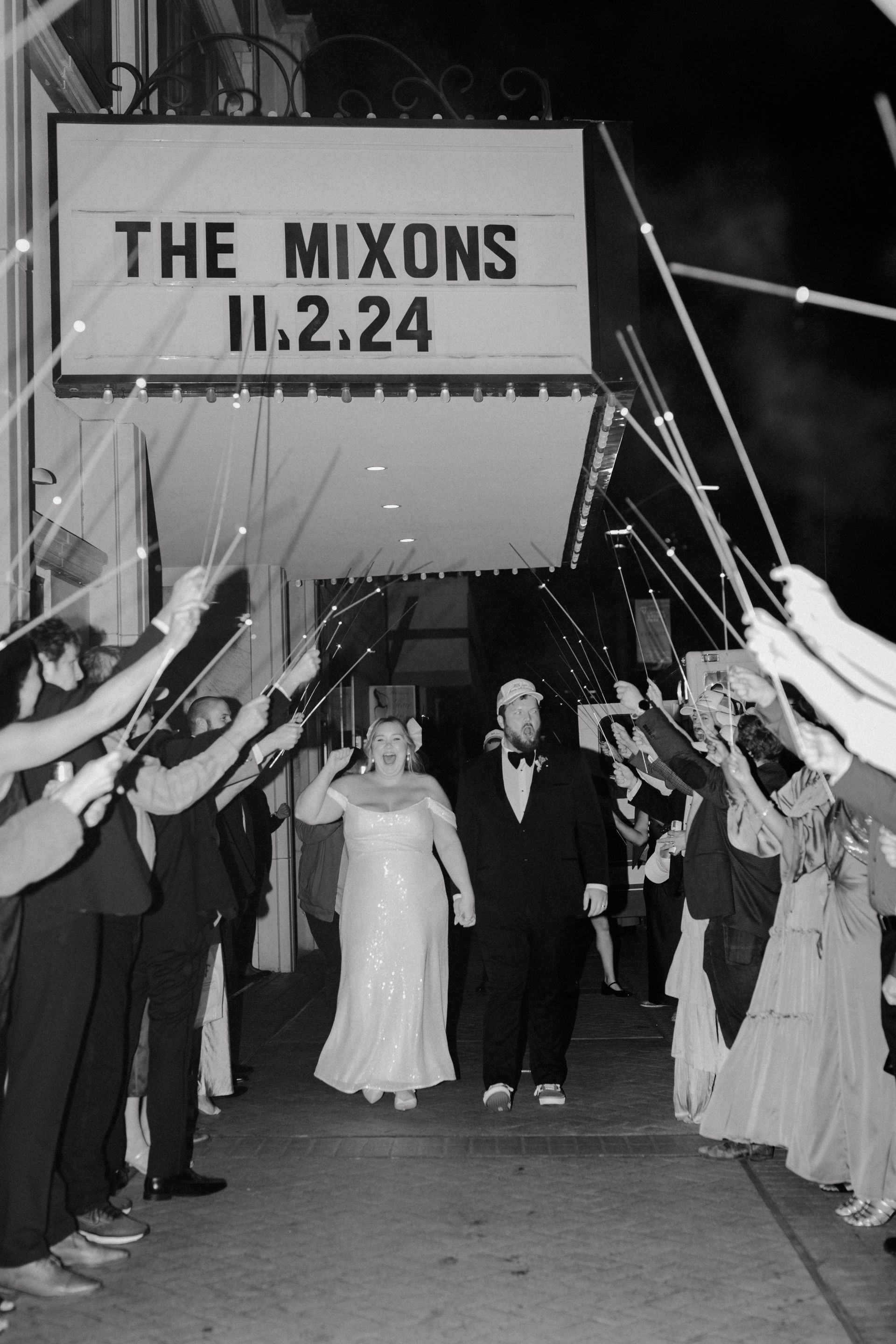 Newlyweds exit venue under sparkler arch; sign reads 