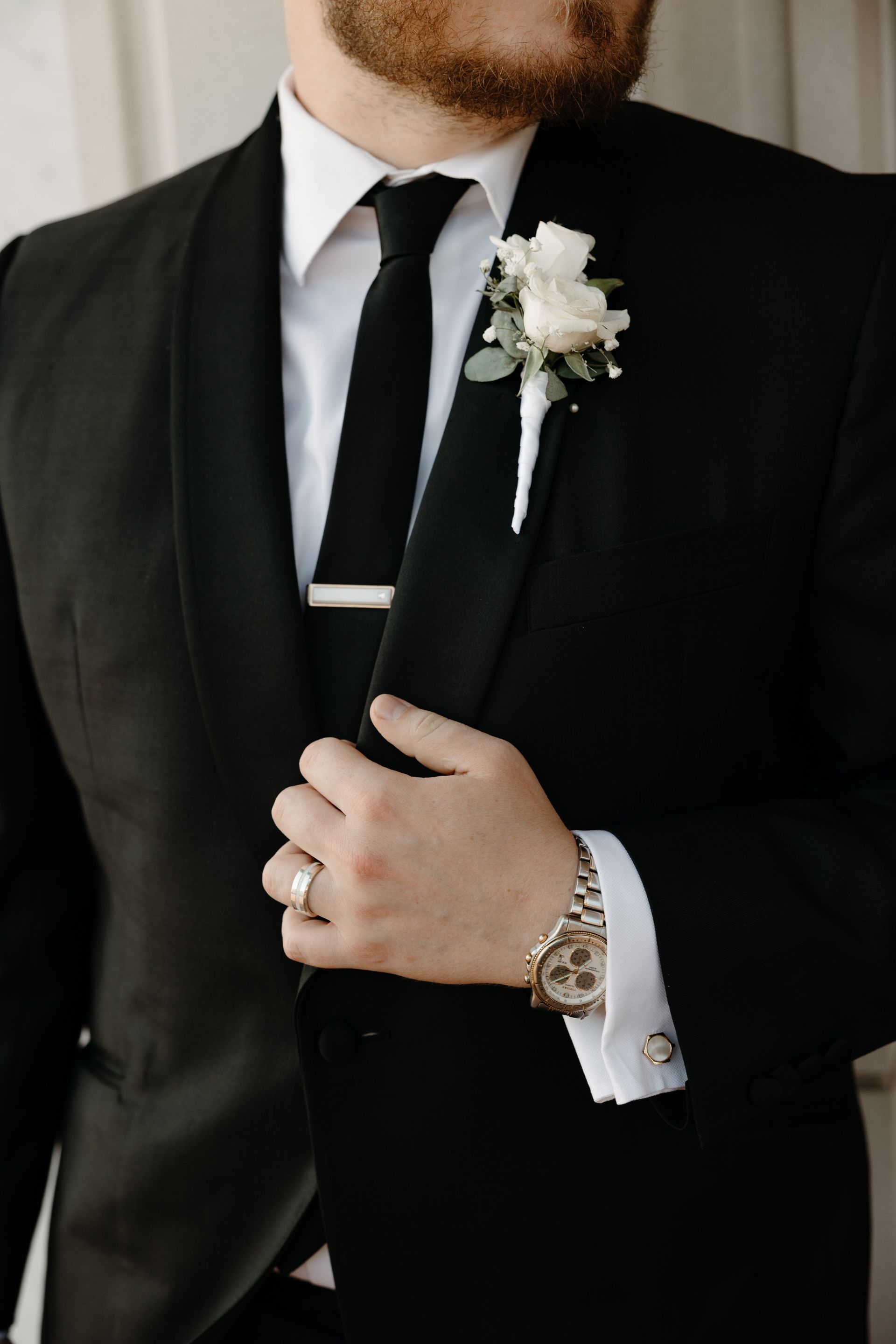 Man in black suit with tie, boutonniere, and watch, adjusting his jacket.