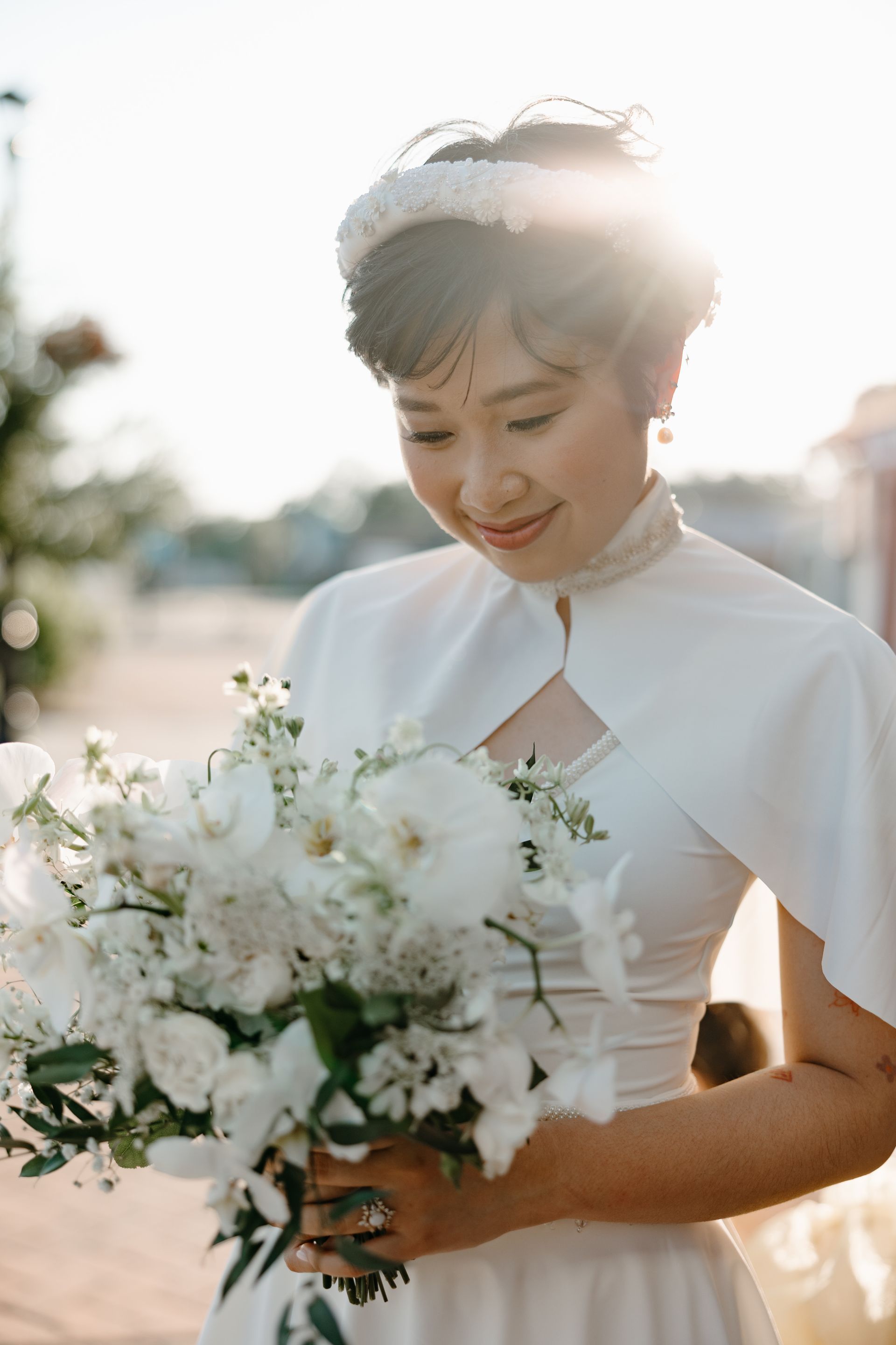 Woman in a white dress and floral headpiece, holding a white bouquet, smiling. Outdoors, sunlit.