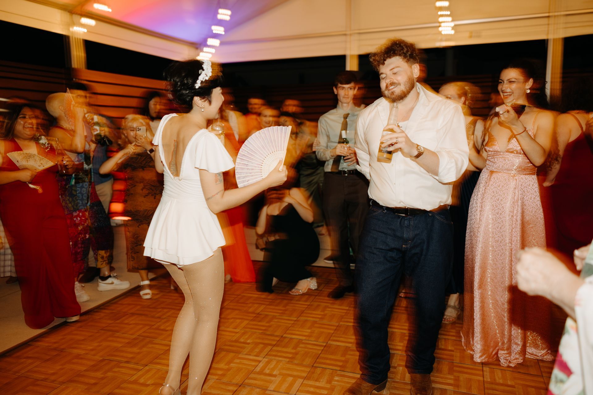 People dancing at an event under a white-lit ceiling. Woman in white outfit waves a fan. Man in button-up holds a drink.