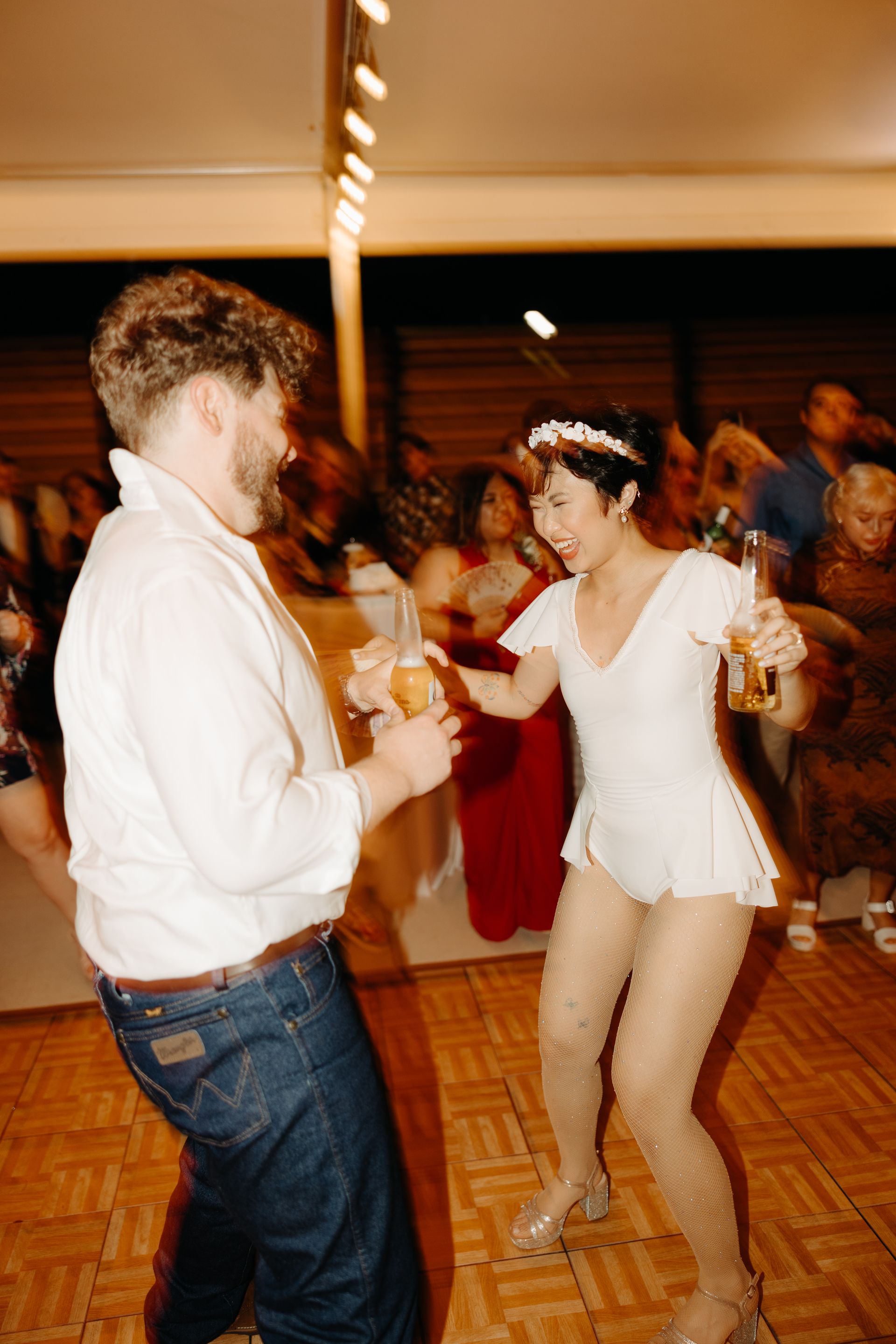 Man and woman dancing, each holding a beer. Woman wears a white one-piece suit. Party setting.