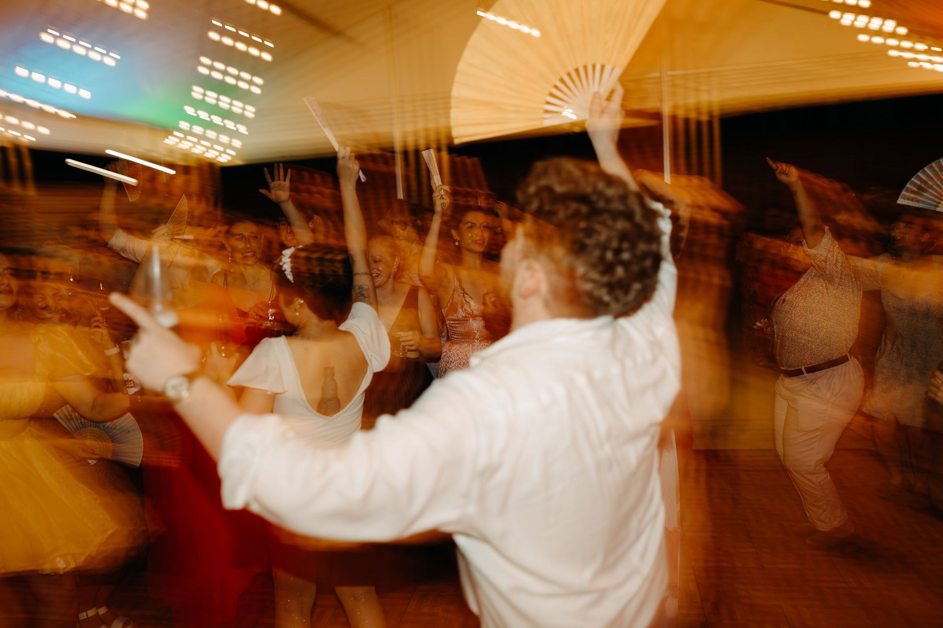 People dancing at a party, some with fans, under bright lights. Blurry motion effect.