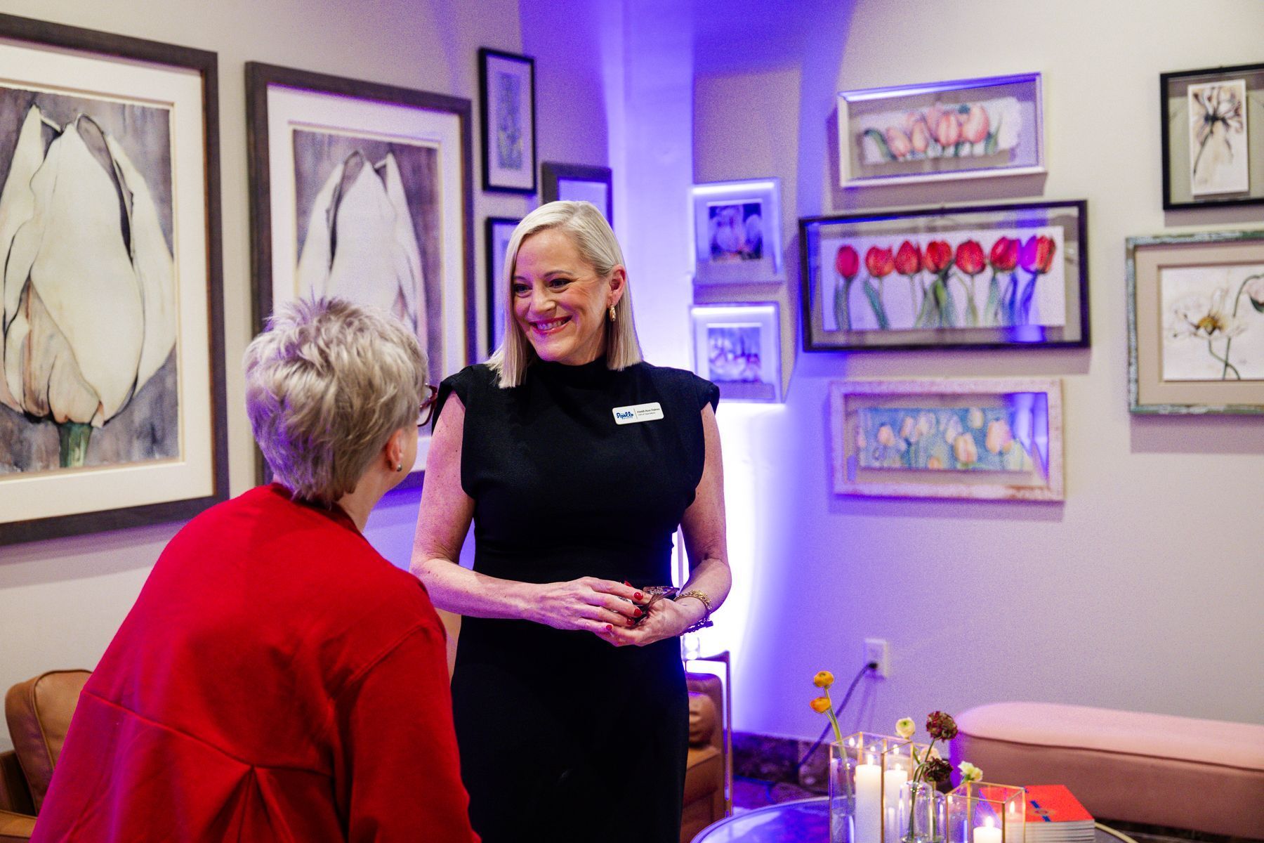 Two women smiling in an art gallery, surrounded by paintings on the wall.