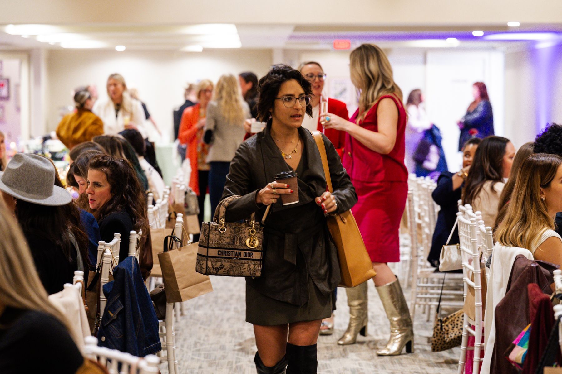 Woman walks through event, holding cup and bag. Other women sit, stand, and chat in room.