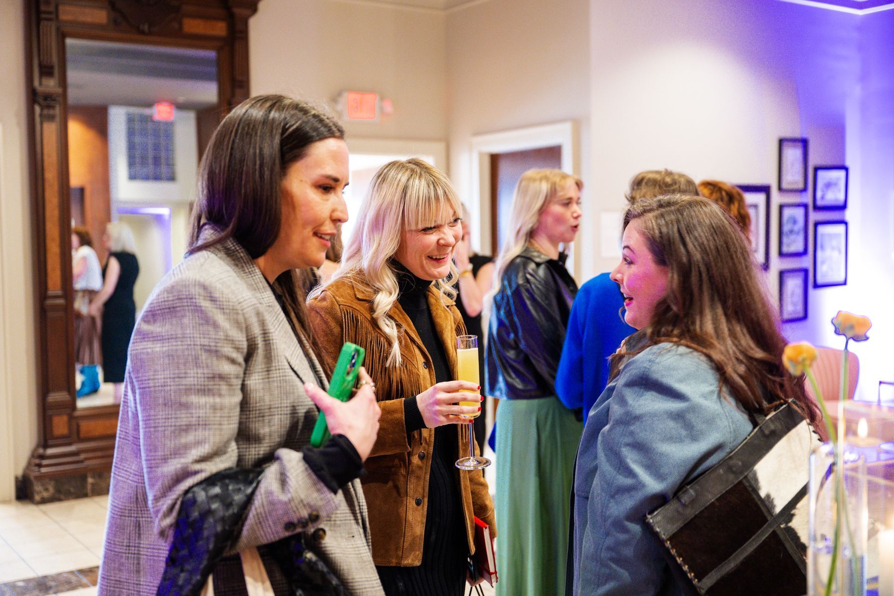People talking at an event; women with drinks and jackets. Interior setting, well-lit with a mirror.