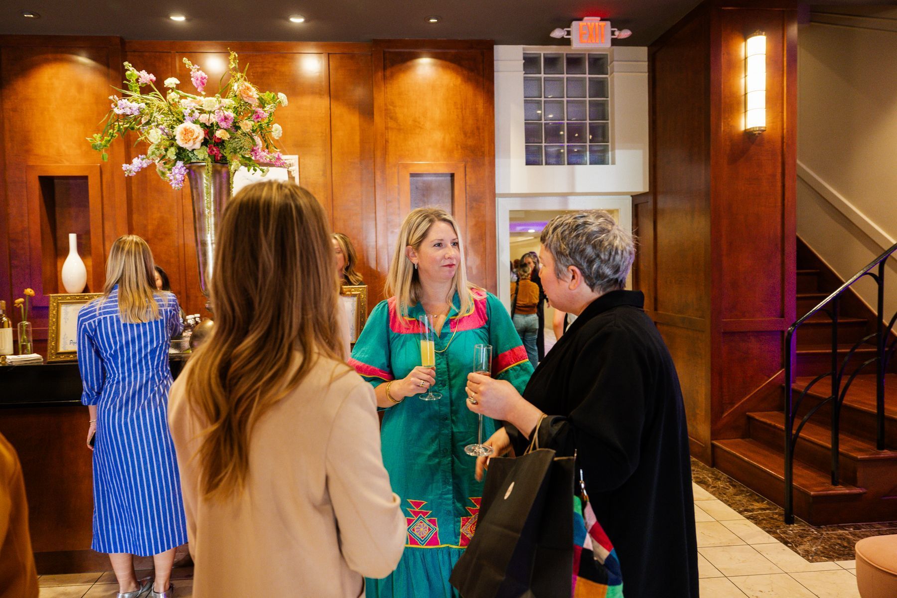 People chatting in a warmly lit interior; woman in teal dress and others with drinks.
