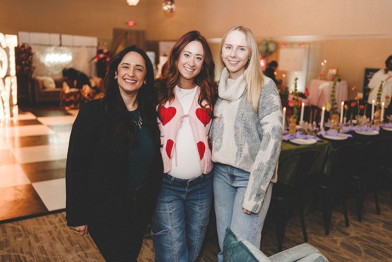 Three women smiling, posing for photo at decorated event with checkered floor and table setting.