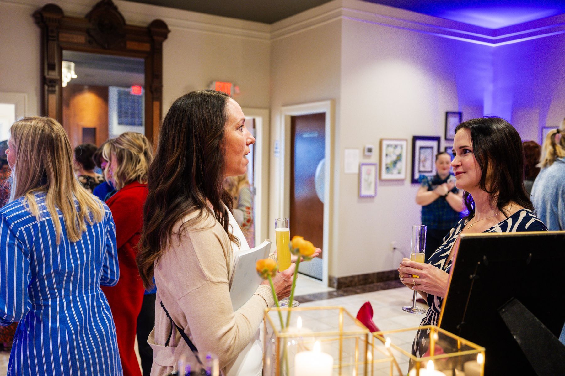 Two women conversing at an event, one holding a drink, with other people in background.