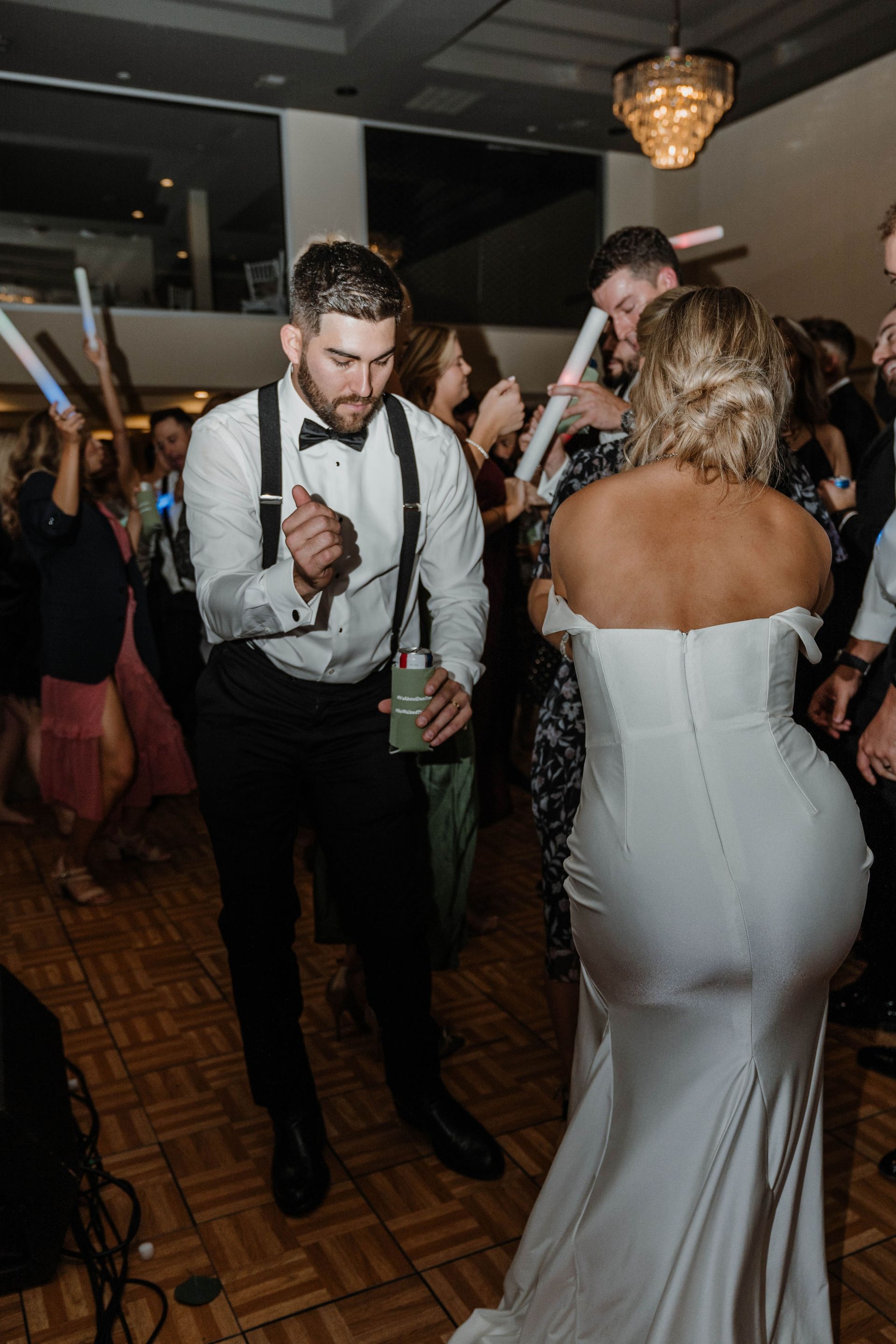 Groom in suspenders dances with bride in white gown at a wedding reception, holding a drink; people with glow sticks in background.