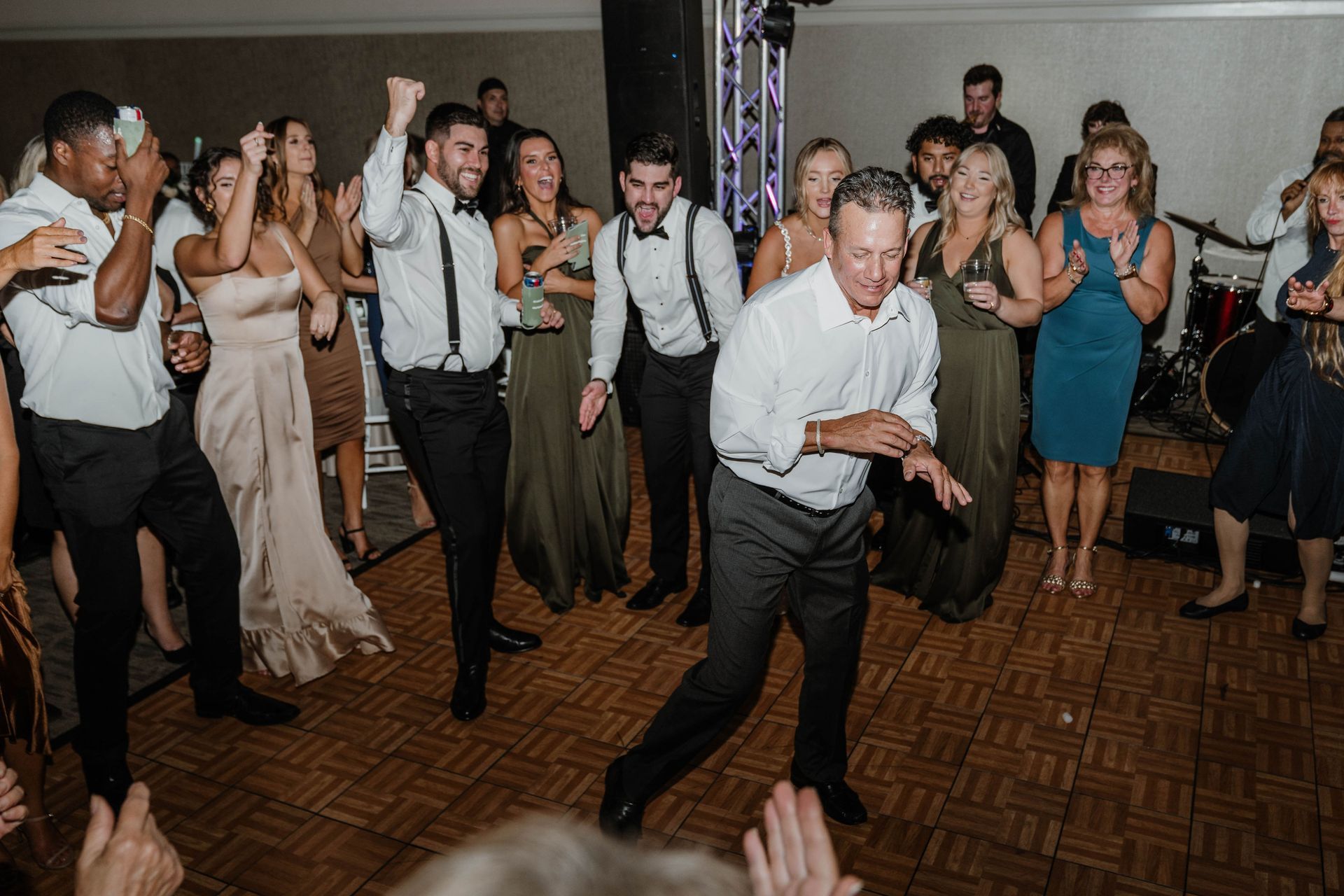 People dancing at a wedding reception. Some are raising their arms, smiling, and clapping. Wooden dance floor.