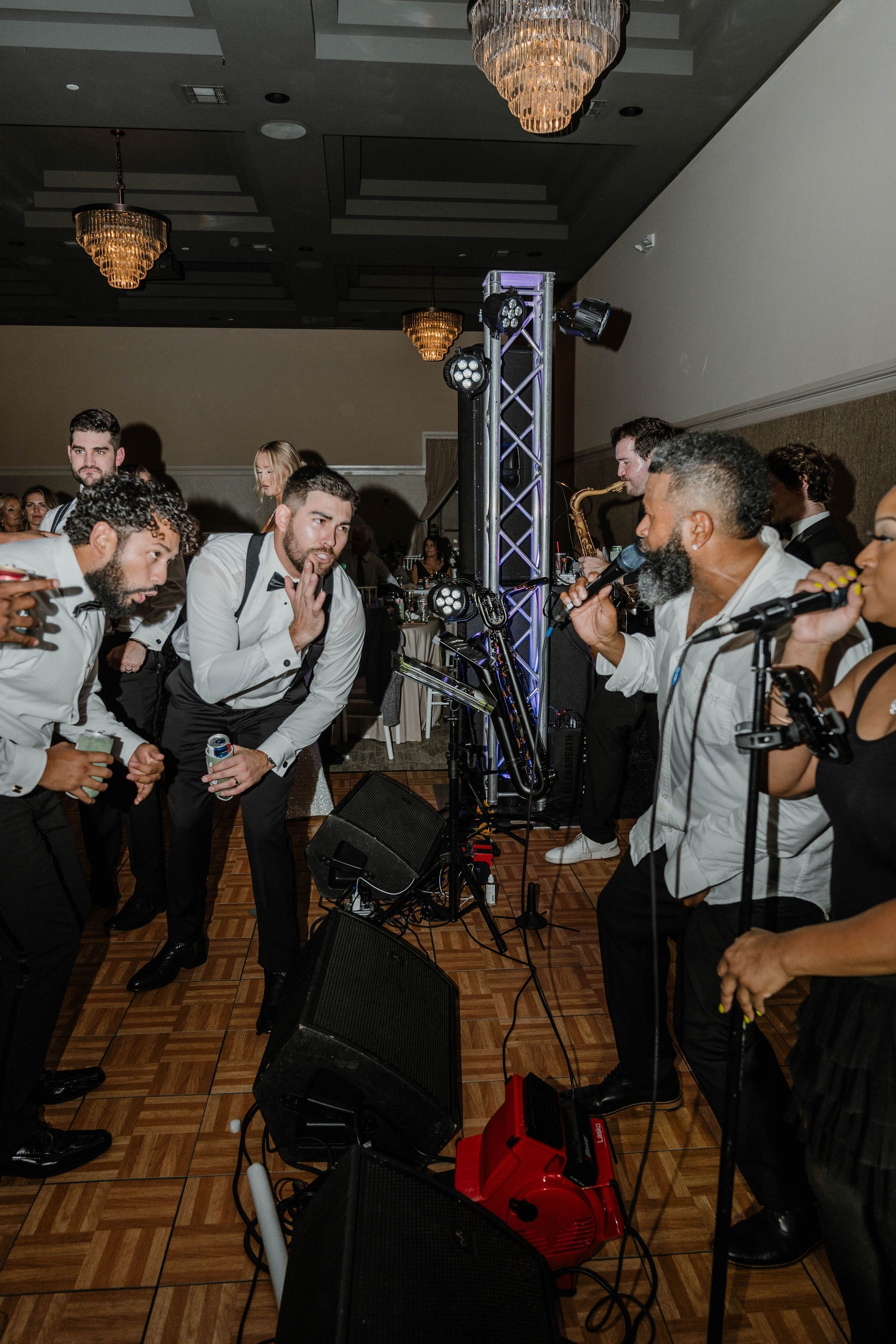 Band performing at an event, some members singing and playing instruments. Dimly lit room with wood floor.