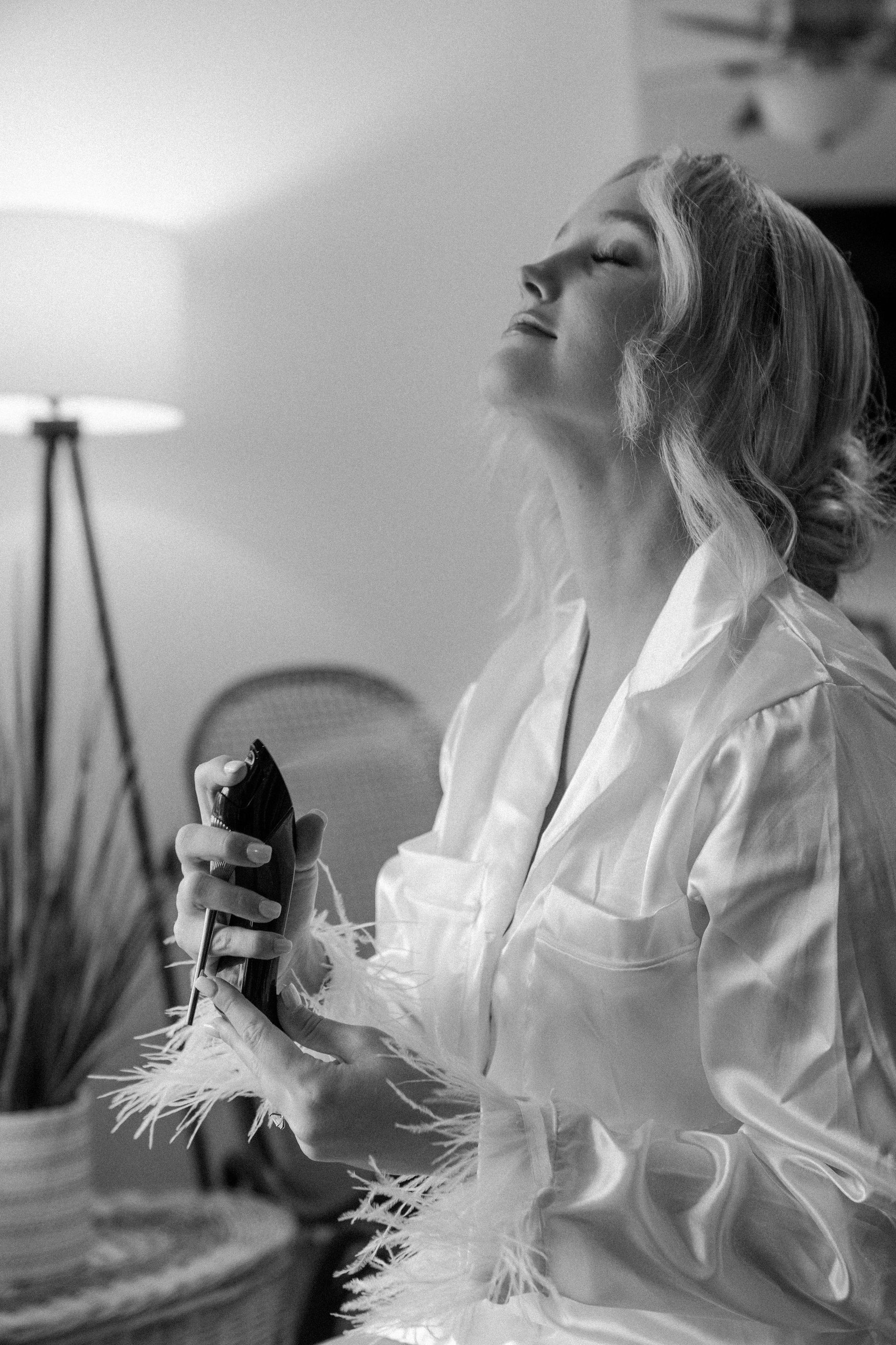 Woman sprays perfume, eyes closed, wearing a white robe with feather trim indoors near a lamp.