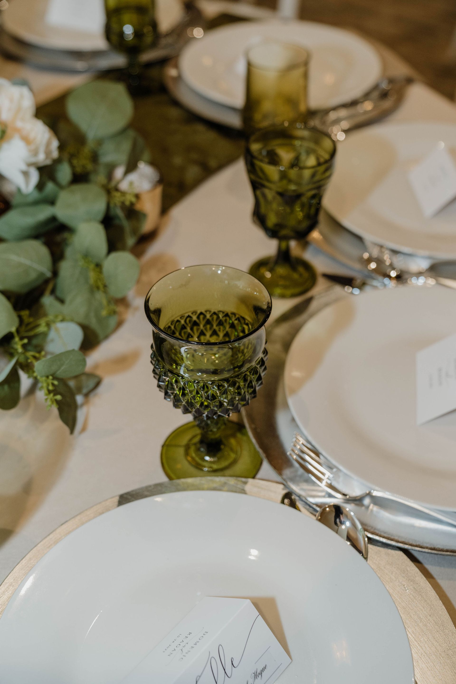 Close-up of a table setting with green goblets, white plates, and silver chargers.
