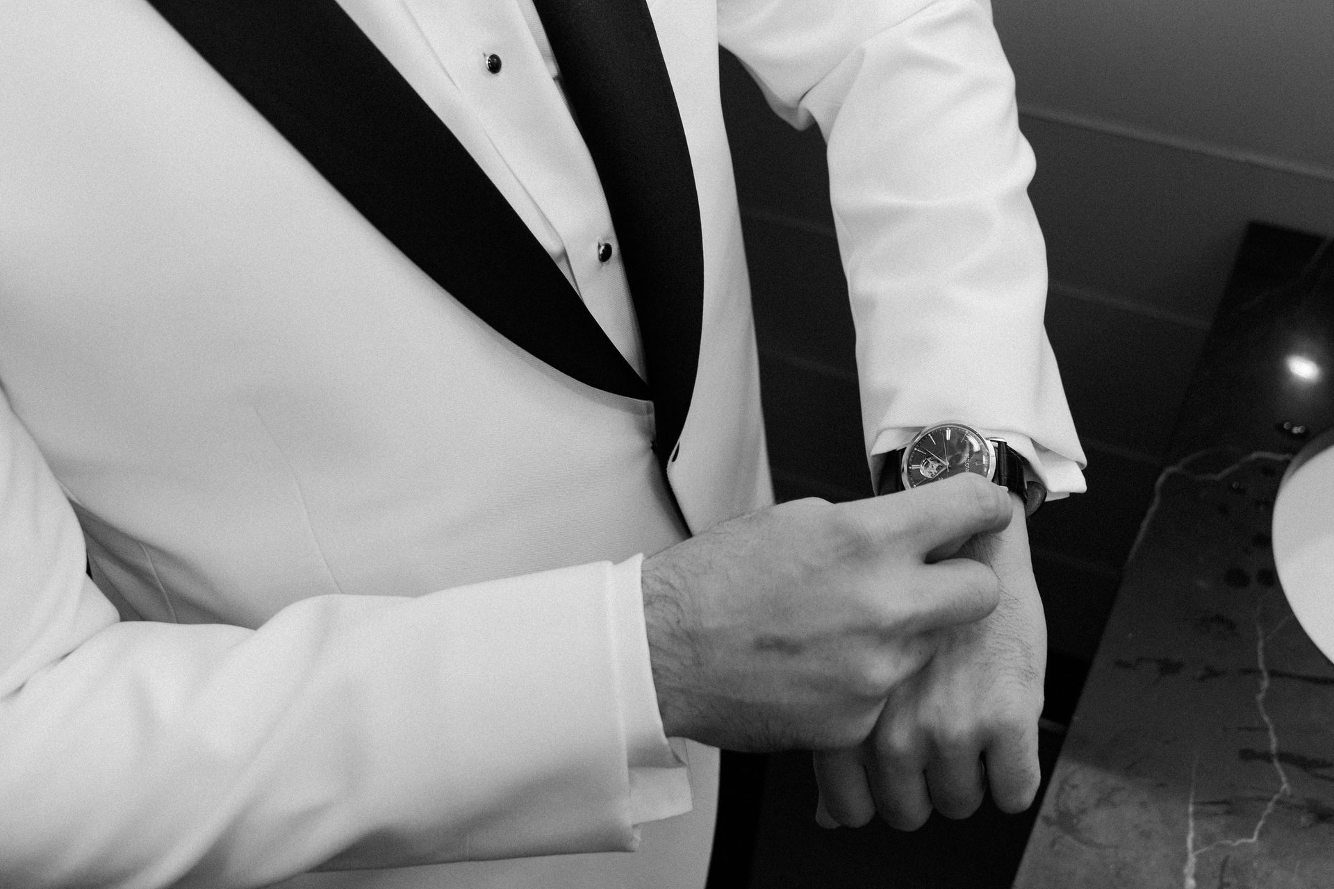 Man in white tuxedo jacket adjusts watch on wrist. Black and white.