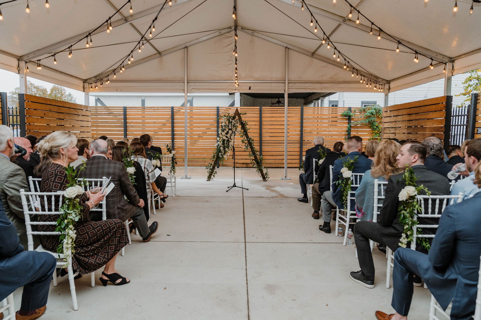 Wedding ceremony in a tent. Guests seated in white chairs, facing an arch decorated with greenery and flowers. String lights hang above.