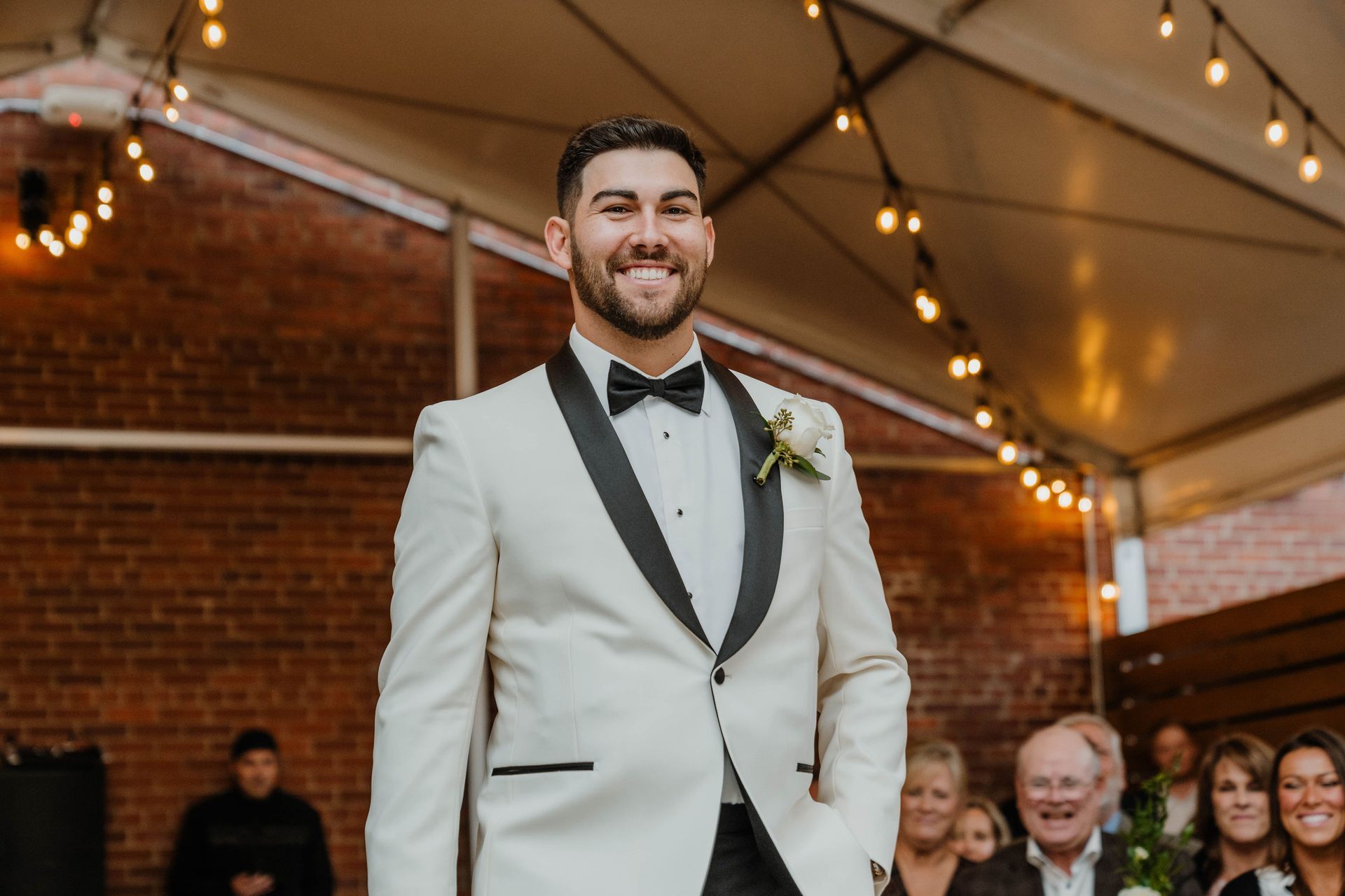Man in cream tuxedo smiles in an outdoor setting with string lights and brick wall.