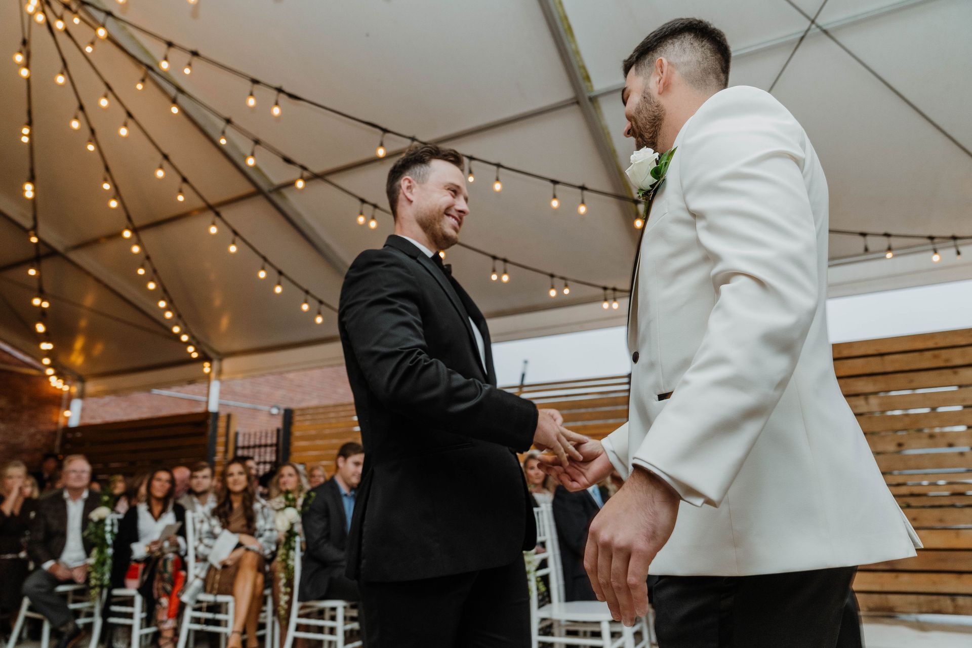 Two men shake hands during an outdoor wedding ceremony, under string lights. Guests watch from seats.