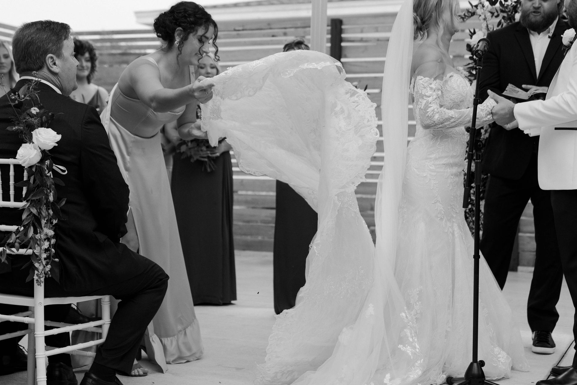 Wedding ceremony: bridesmaid adjusts bride's veil. Groom sits, officiant and another guest stand nearby. Outdoors.