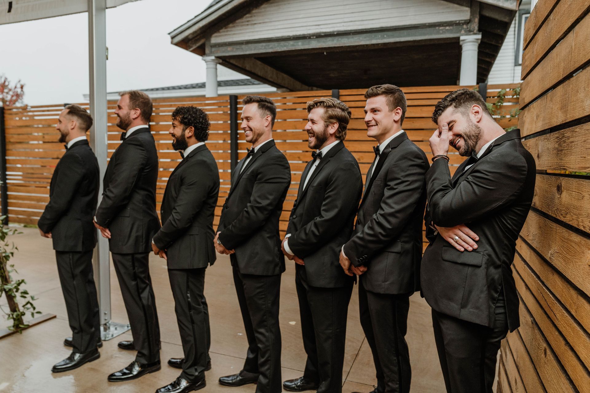 Men in black tuxedos laugh outdoors near a wooden wall, anticipating a wedding.