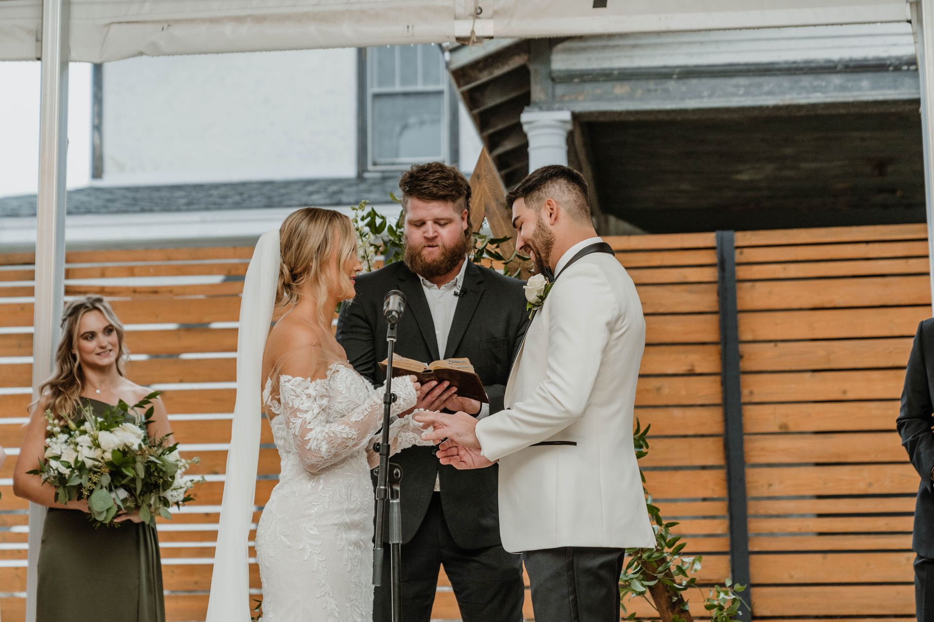 Wedding ceremony. Bride and groom exchange rings, officiant reads. Bridesmaid observes. Wooden backdrop.