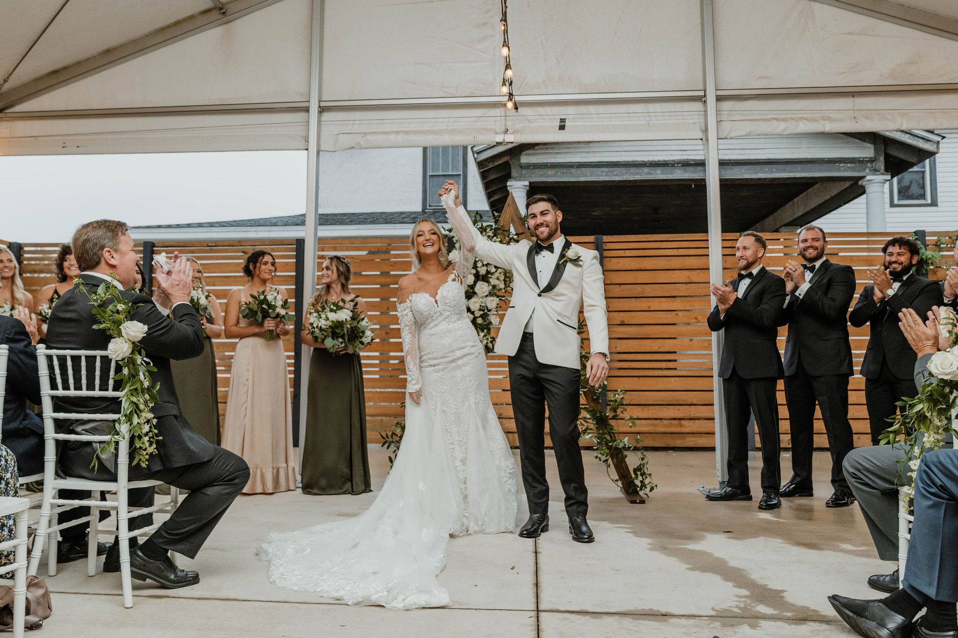 Newlyweds celebrate, raising arms, under a tent, with cheering wedding guests.