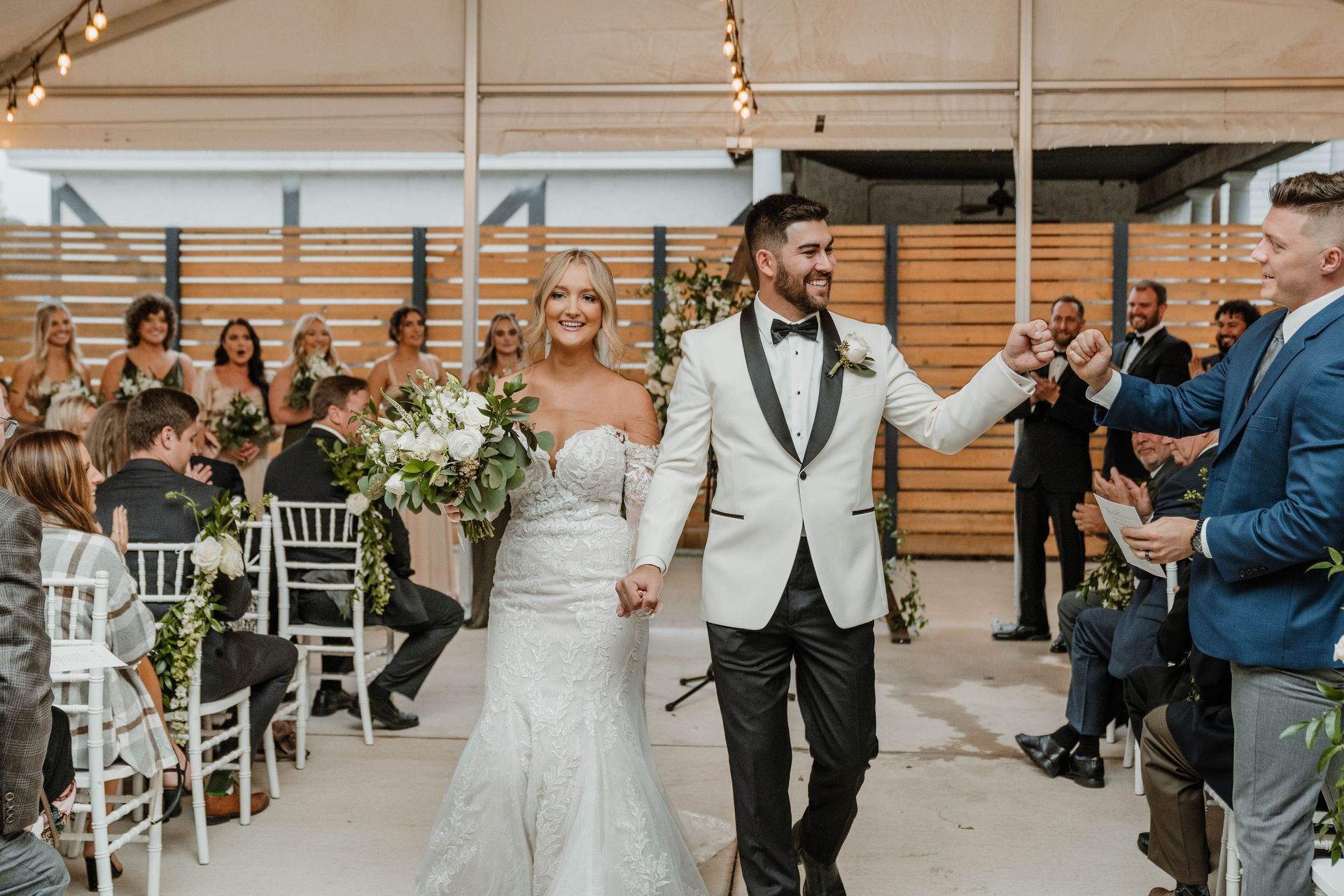 Newlyweds walking down the aisle, holding hands. Bride in white dress, groom in black and white suit. Guests seated, cheering.